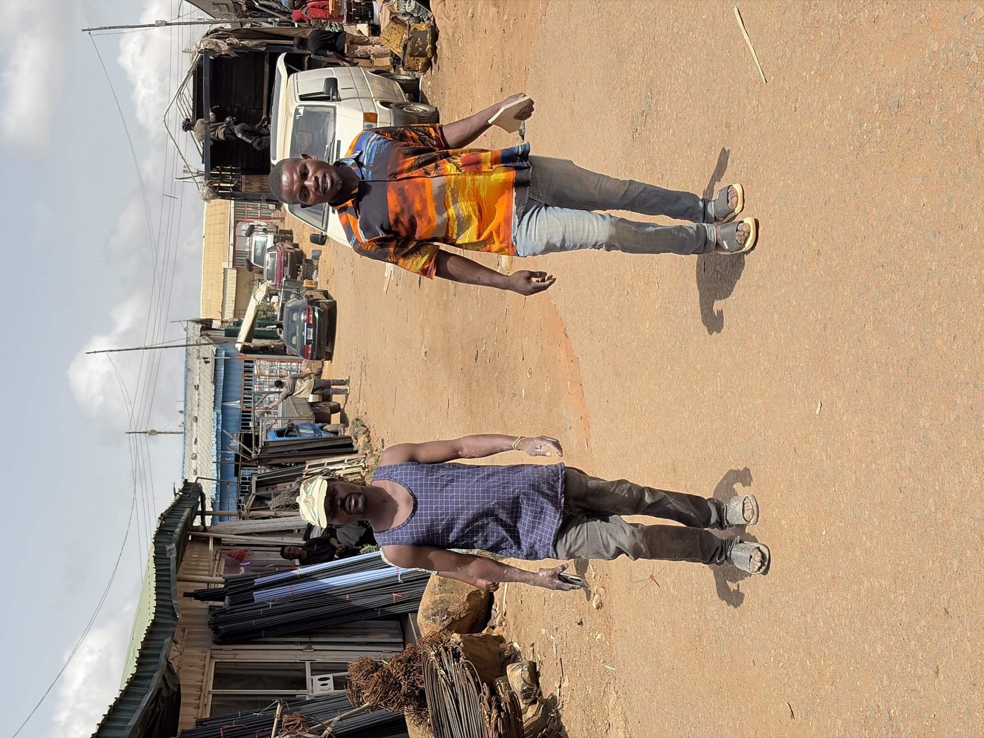 The image depicts an outdoor scene in Gura Top, Nigeria. Two men stand on a dirt road. One man wears a colorful button-down shirt, jeans, and sandals. The other man wears a blue tank top, dark pants, a baseball cap, and sandals. Buildings and stacked metal rods appear in the background. A white truck is also in view.
