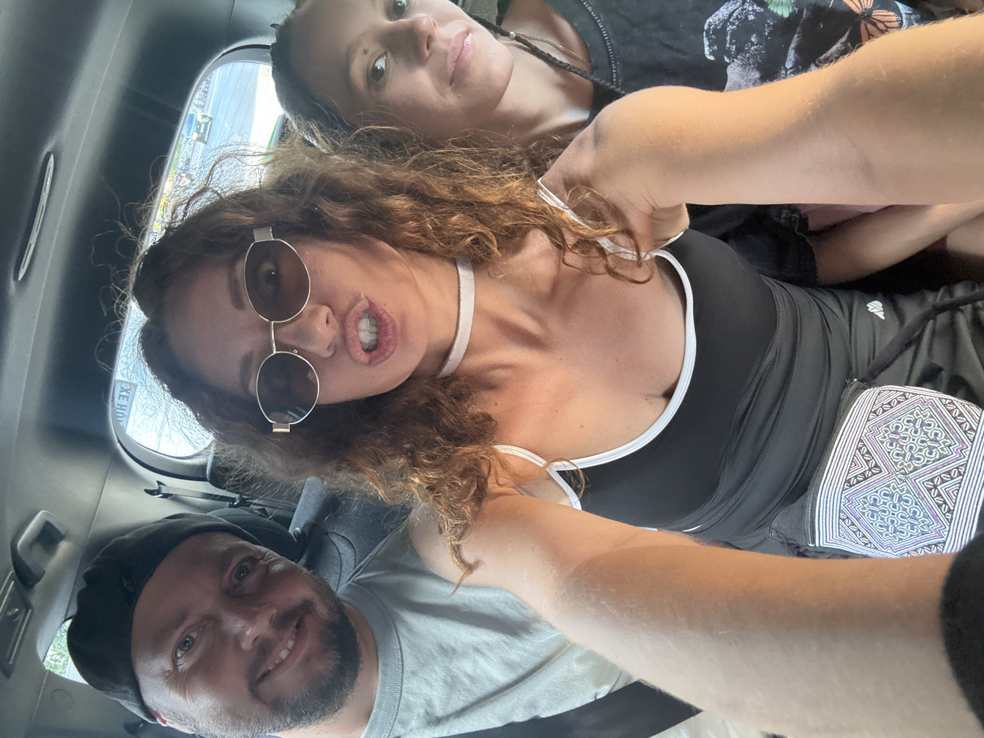 Three people are inside a vehicle, taking a selfie. On the left, a man wearing a black beanie and a light-colored t-shirt smiles at the camera. In the center, a woman with curly brown hair and sunglasses makes a duck face. She is wearing a black tank top with a white trim and a white choker necklace. To the right, another woman, partially visible, looks towards the camera with a neutral expression. Her dark hair is braided. The interior of the vehicle is visible, with the rear window showing an outdoor scene with trees and what appears to be a road with a car. The image appears to be taken in Hà Nội, Vietnam, given the context of previous interactions.