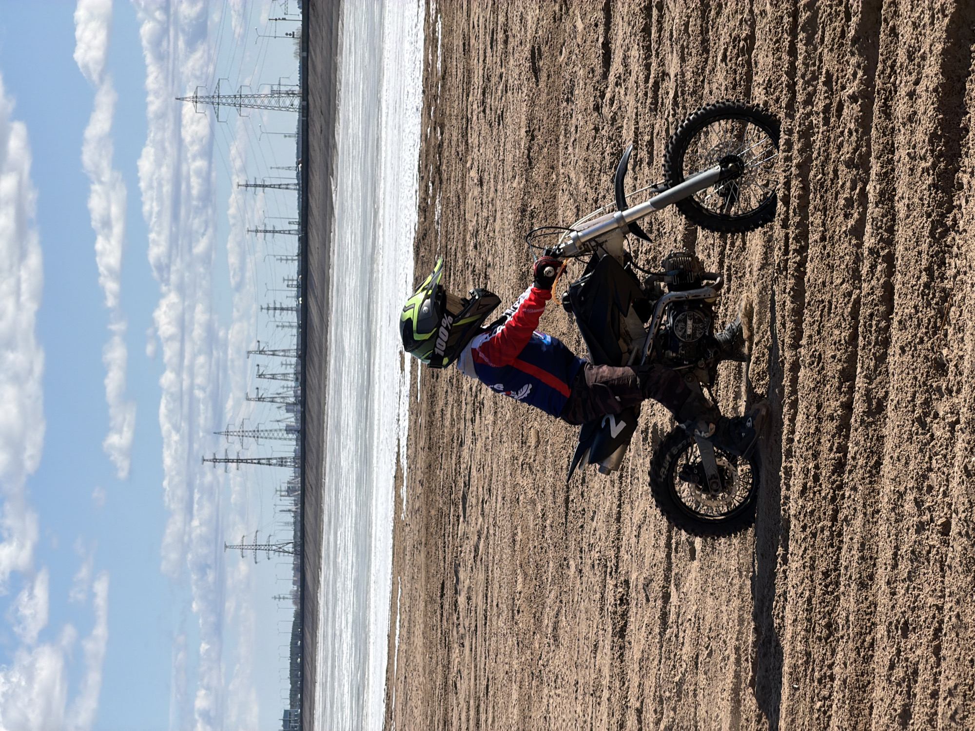 A man is riding a small dirt bike on a sandy or muddy area. He is wearing a helmet, red and blue riding gear, and black boots. The dirt bike is small and appears to be a minibike or pit bike. In the background, there is a large body of water or a frozen area, with power lines visible in the distance. The scene appears to be outdoors in Chaikovsky, Russia.
