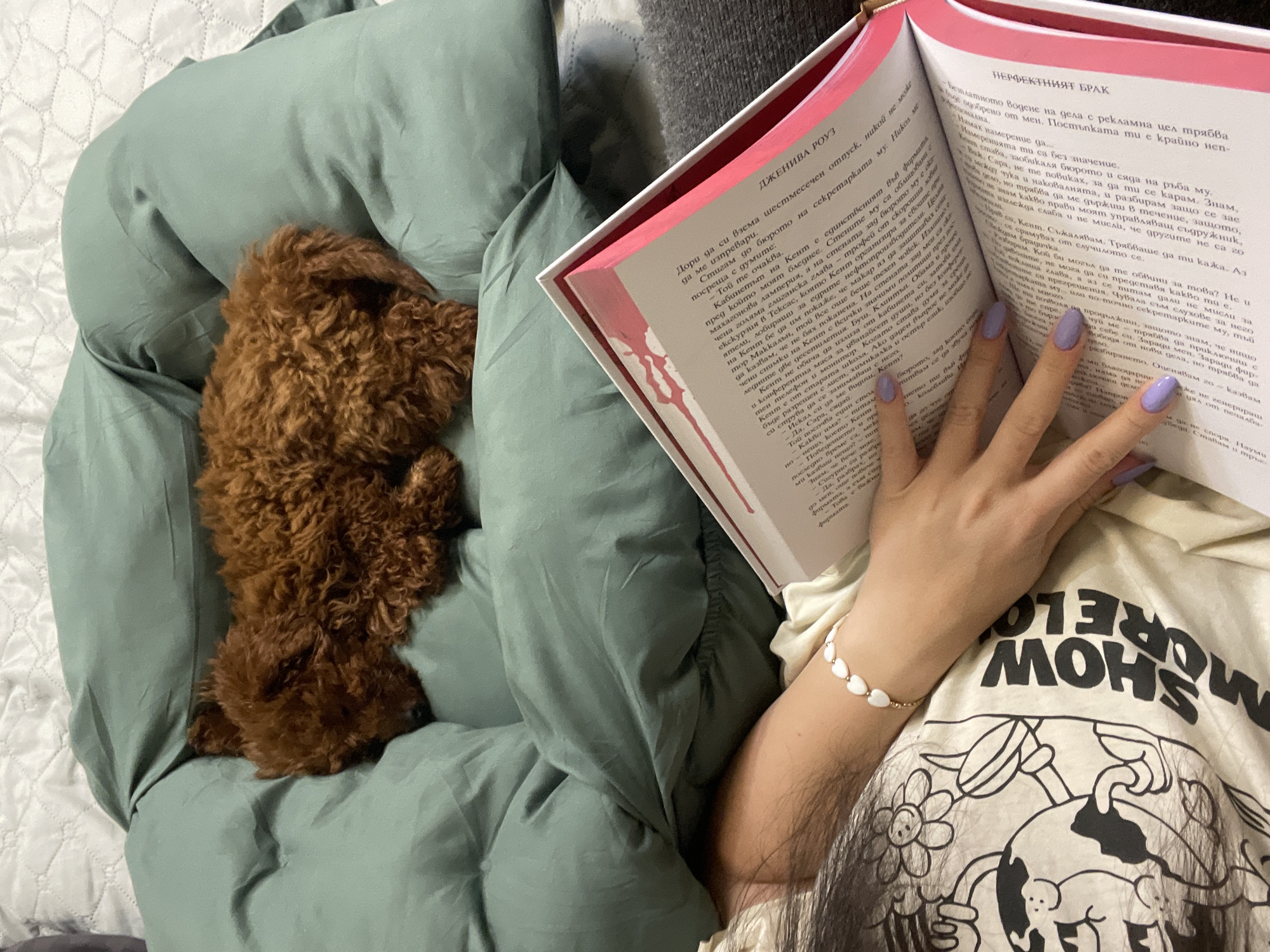 A person is reading a book in Bulgarian in bed. The book, partially visible, is titled "The Perfect Marriage". The person is wearing a light-colored t-shirt with a graphic of a globe with cartoonish figures and the words "SHOW MORE LOVE" written on it. They also have a bracelet of small white hearts on their left wrist and lilac-colored nail polish. A small, brown poodle is lying near them on the green bedding. The location is most likely in a private residence in Sofia, Bulgaria, given the Bulgarian language book.