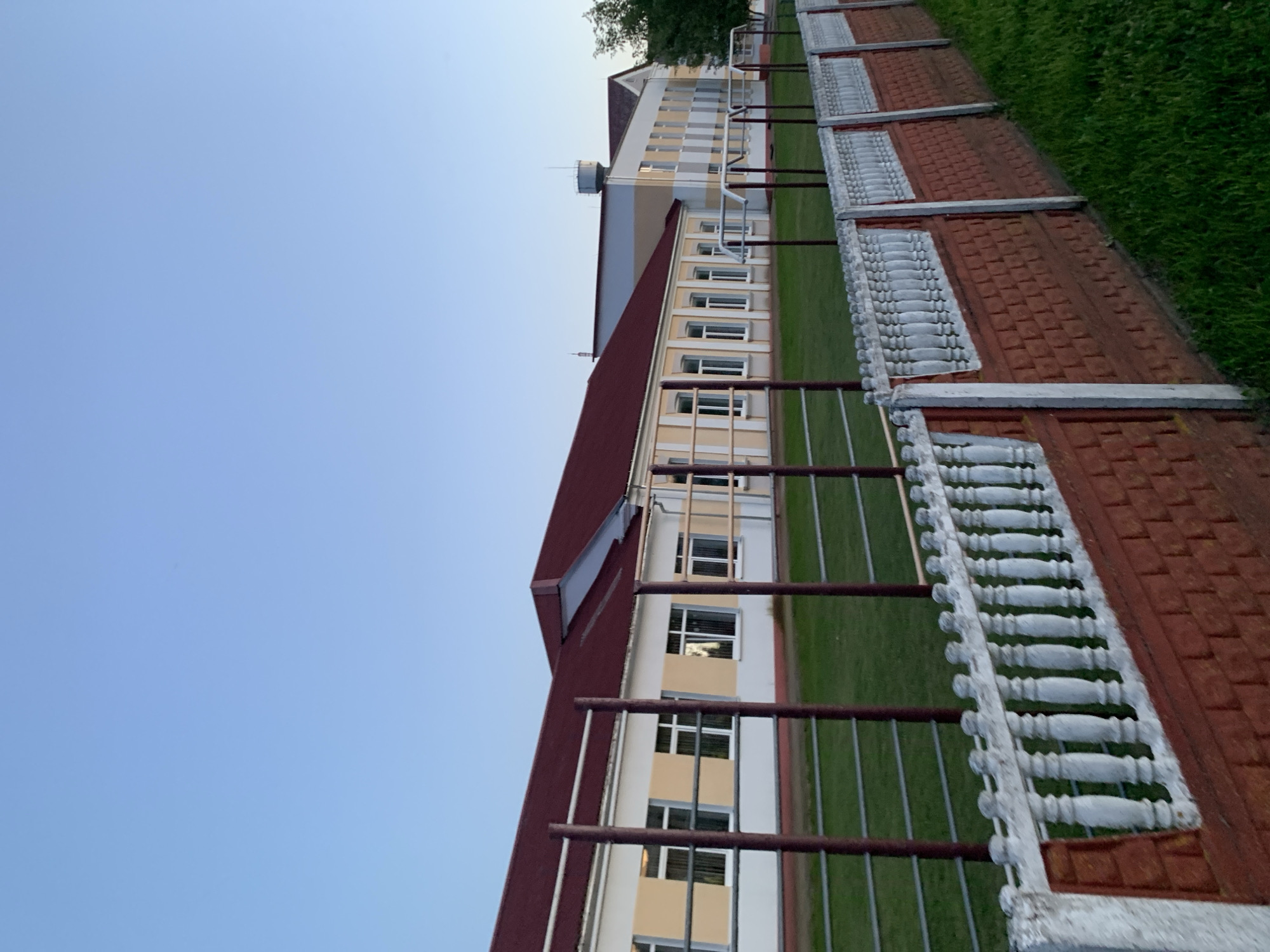 The image shows a building with a red roof and yellow and white walls, possibly a school or community center, in Kapyĺ, Belarus. In front of the building is a grassy field with some sports equipment, such as horizontal bars. A red brick fence with white decorative elements runs along the front of the property, separating the field from a grassy area. The sky is clear and blue, suggesting it is daytime. There are no people visible in the image.