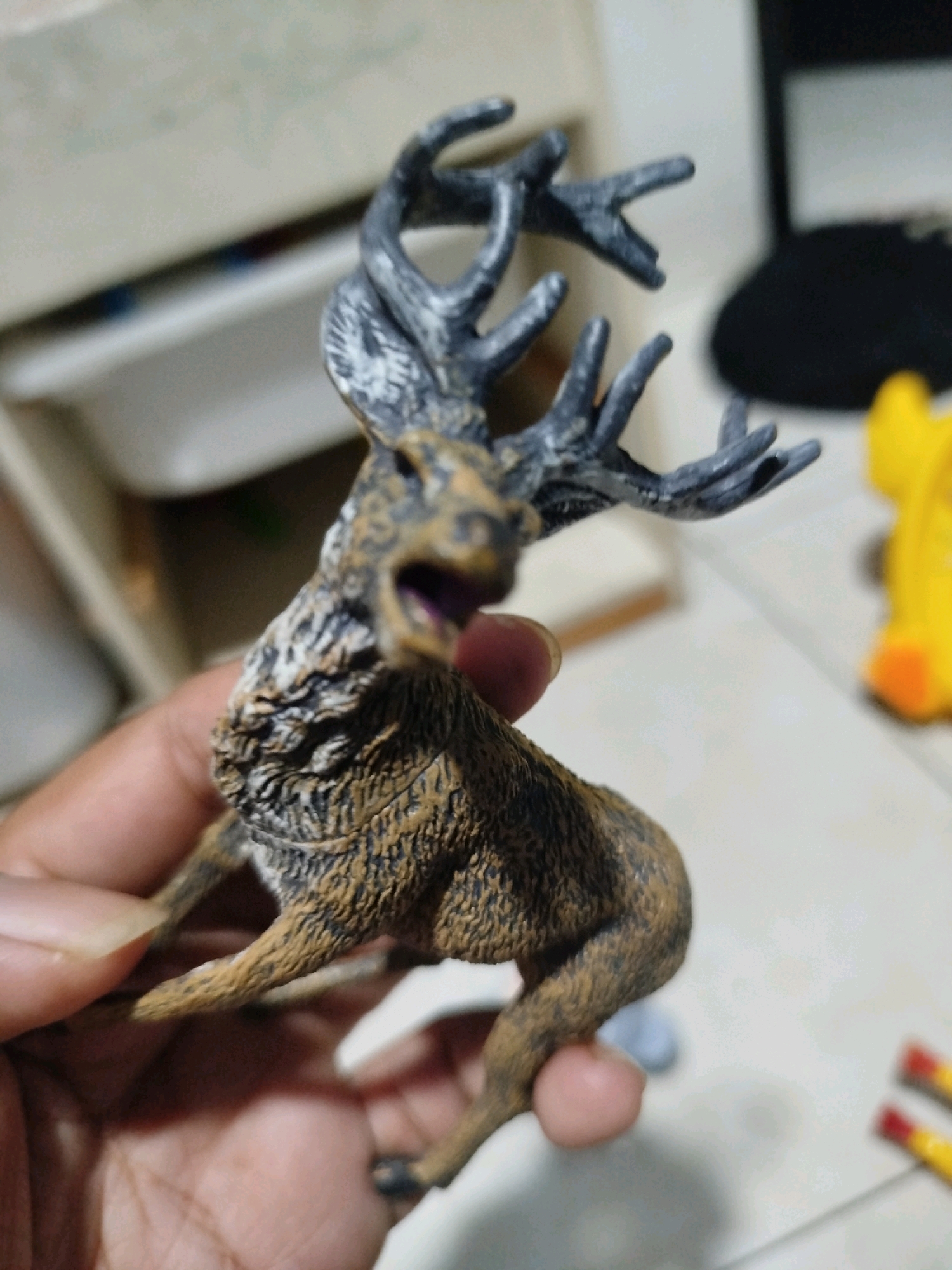 A close-up, slightly blurred, medium shot shows a hand holding a detailed, brown and grey toy deer with large antlers. The deer is facing left with its mouth open, appearing to be in mid-stride or mid-action. The background is out of focus, with hints of indoor furniture and objects, including a blurry yellow toy in the upper right corner and two colorful objects resembling pens or pencils on the right edge of the frame. The lighting is ambient and appears to be artificial, casting soft shadows. The scene is an indoor setting, possibly a home or office, and no specific location in Mojoagung, Indonesia, is discernible from the visual evidence.