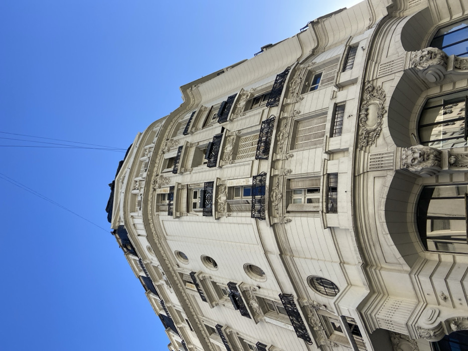 An ornate, light-colored building angles against a clear blue sky. Windows of varying sizes and shapes are visible, some with dark decorative ironwork. Architectural details include protruding stone carvings and rounded window arches. Thin wires cut across the upper left of the frame, creating a stark contrast against the classic facade. The building curves, presenting a unique perspective.