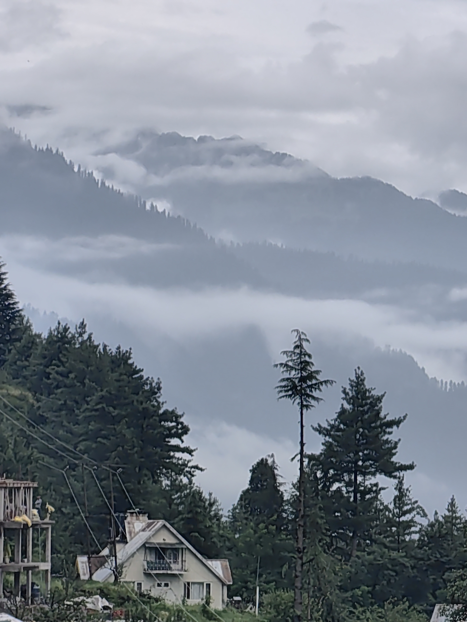 The image captures a mountainous landscape with a hazy, overcast sky. In the foreground, a cluster of evergreen trees is visible, with some of them tall and slender. To the left of the trees, a partially constructed concrete building stands, appearing unfinished. To the right of the trees, a light-colored house with a sloped roof and a chimney is situated on a hillside, surrounded by foliage. The middle ground features layers of misty mountains receding into the distance. Electrical wires are strung across the scene, connecting to poles. The overall impression is one of a rural, mountainous setting, possibly in or near Manali, India, given the distinctive architecture and mountainous terrain.