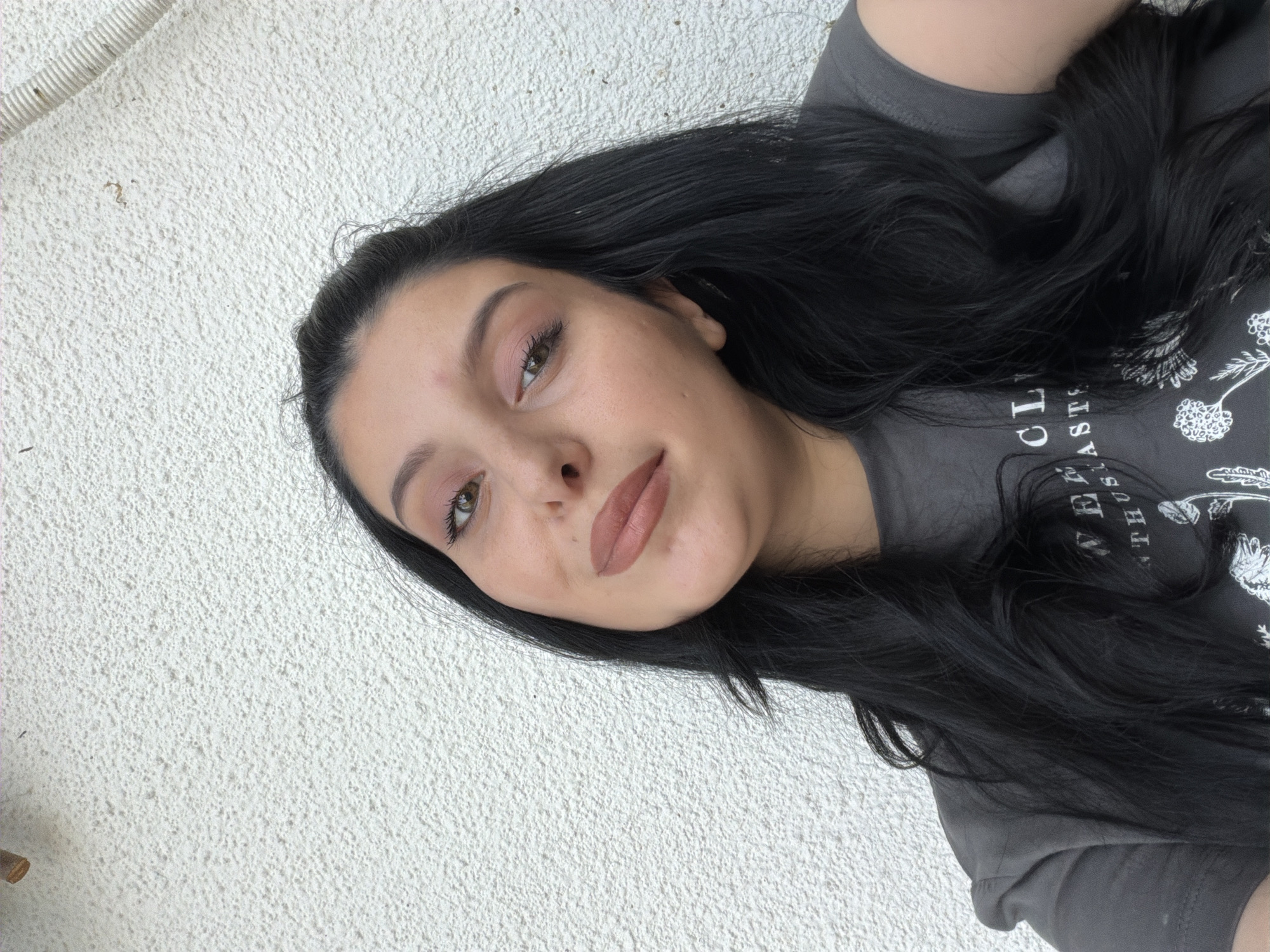 A young woman with long, dark hair is pictured in a close-up shot against a textured white wall. She is wearing a dark gray t-shirt with a graphic design. Her head is tilted slightly, and she is looking towards the camera with a neutral expression. The lighting suggests an outdoor setting, possibly during the daytime. The image does not contain any information that would allow for the determination of its location in Tbilisi, Georgia, or any other specific city.