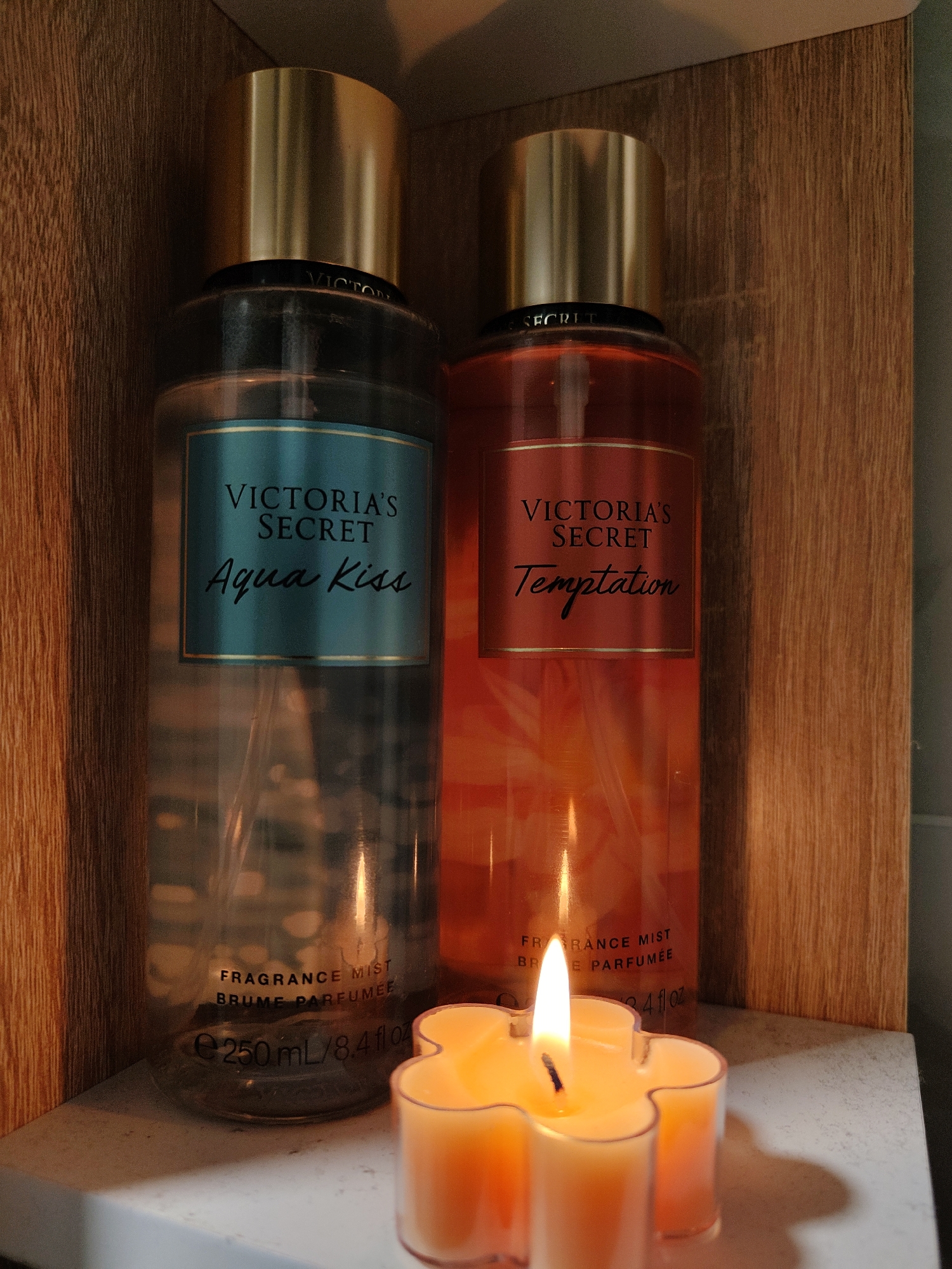 The image shows two bottles of Victoria's Secret fragrance mist placed on a shelf next to a lit flower-shaped candle. The bottle on the left is "Aqua Kiss", and the one on the right is "Temptation." Both bottles have gold caps. The background consists of wooden panels, suggesting the shelf is part of a cabinet or closet. The candle is orange and yellow and casts light on the bottles. The image appears to have been taken in an indoor setting. There is no information in the image to suggest this scene is in Moscow, Russia.