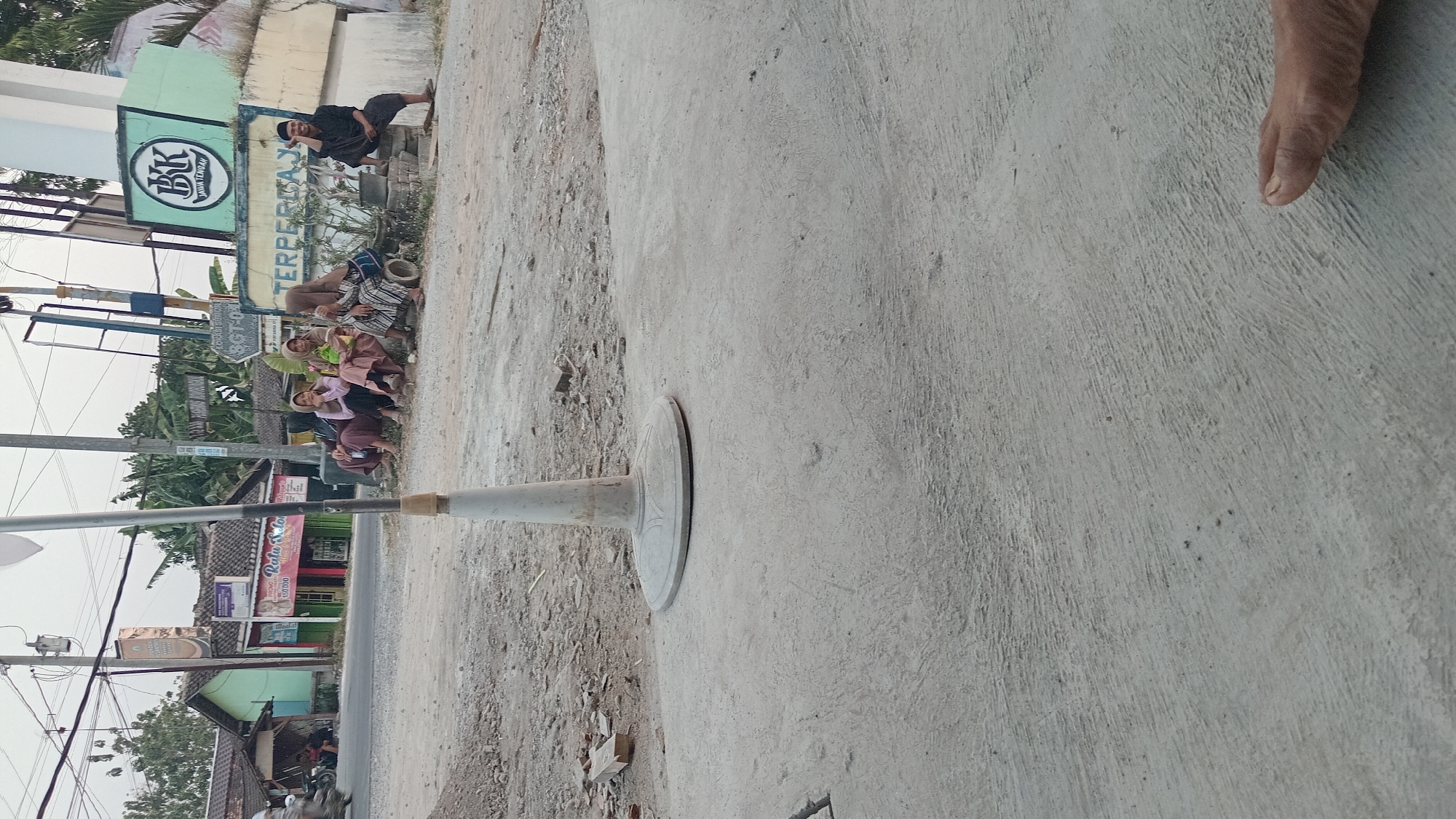 The image depicts a street scene in Trembulrejo, Indonesia, with a focus on a group of people gathered by the side of the road. In the foreground, a concrete surface with some rough texture is visible, and a portion of a bare foot is present in the bottom right corner. A white traffic cone, or similar object, stands on a circular base, partially obscuring the view of the background.

In the mid-ground, a dirt and gravel area lines the road. Several people are seated on the ground, appearing to be conversing. Four individuals, likely women, are dressed in traditional Islamic clothing, including headscarves, and are sitting together. To their right, a man in a dark shirt and trousers is seated, looking towards the left and making a gesture with his hand.

Behind the seated individuals, there are several buildings. One building features a sign with "BK JAWA TENGAH" in a logo and text. Another sign in yellow reads "TERPERCAYA". Adjacent to this is a smaller green building with a red and white sign that says "Ratu Salon" and "PROMO 150.000". A motorbike is visible on the left side of the road, suggesting it is a public thoroughfare. The overall environment appears to be an outdoor, semi-urban setting with a mix of commercial and residential structures. The lighting suggests it is daytime, possibly late afternoon or early evening, given the soft light and long shadows.