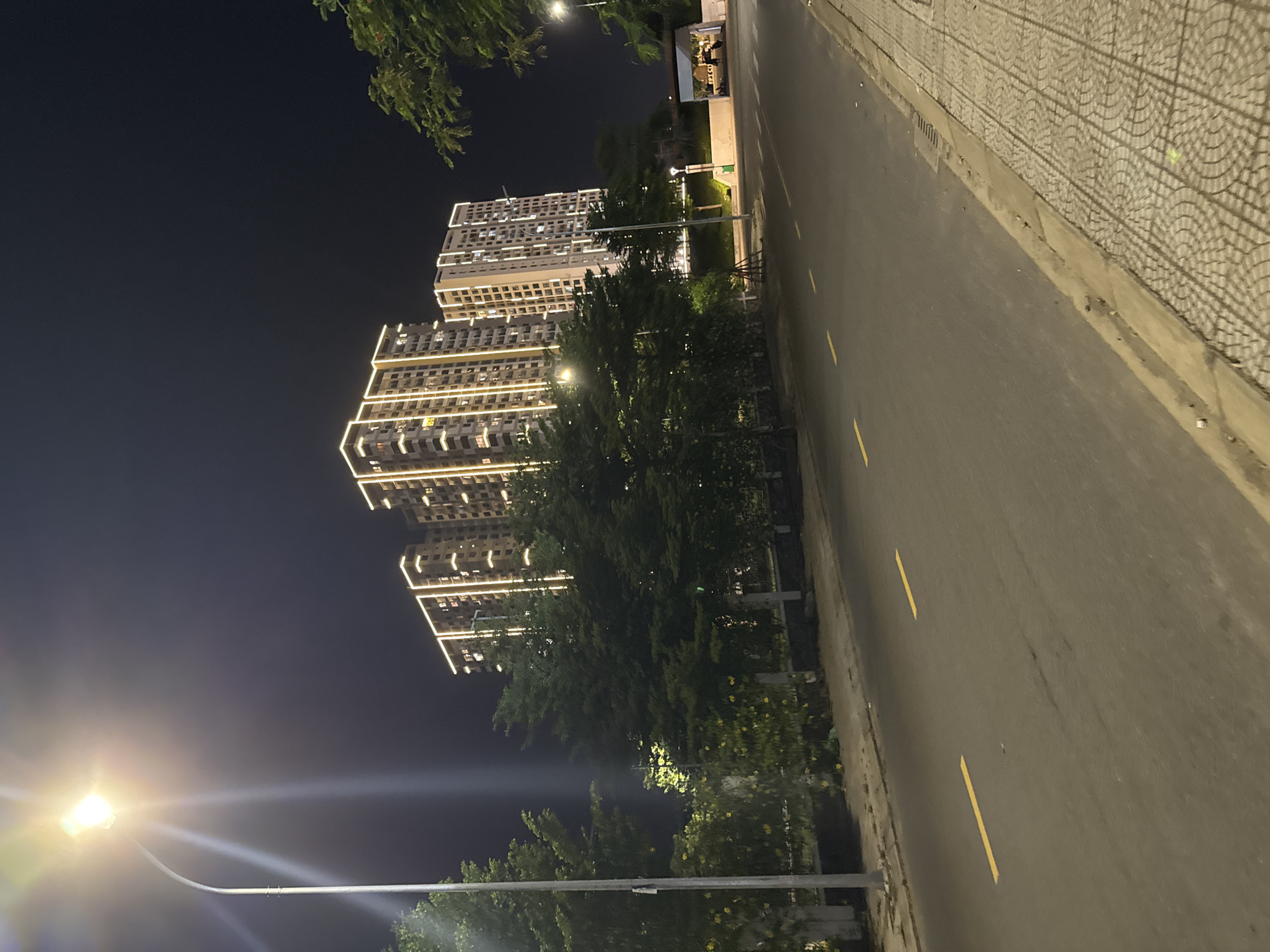 The image depicts a street scene at night in the city of Đà Nẵng, Vietnam, specifically in the vicinity of the Four Points by Sheraton hotel, likely along the coastal road. In the background, two high-rise apartment buildings are illuminated with a decorative lighting scheme that outlines their edges. The buildings are modern in design, with many windows visible. A street lamp with a bright light source is prominent in the left foreground, casting a glare across the image. The street itself is paved and has yellow lane markings. A sidewalk with a patterned paving stone design runs along the right side of the street. Trees with dense foliage line the area between the street and the apartment buildings. The overall environment suggests an urban setting with residential or mixed-use development. There are no people clearly visible in the scene.