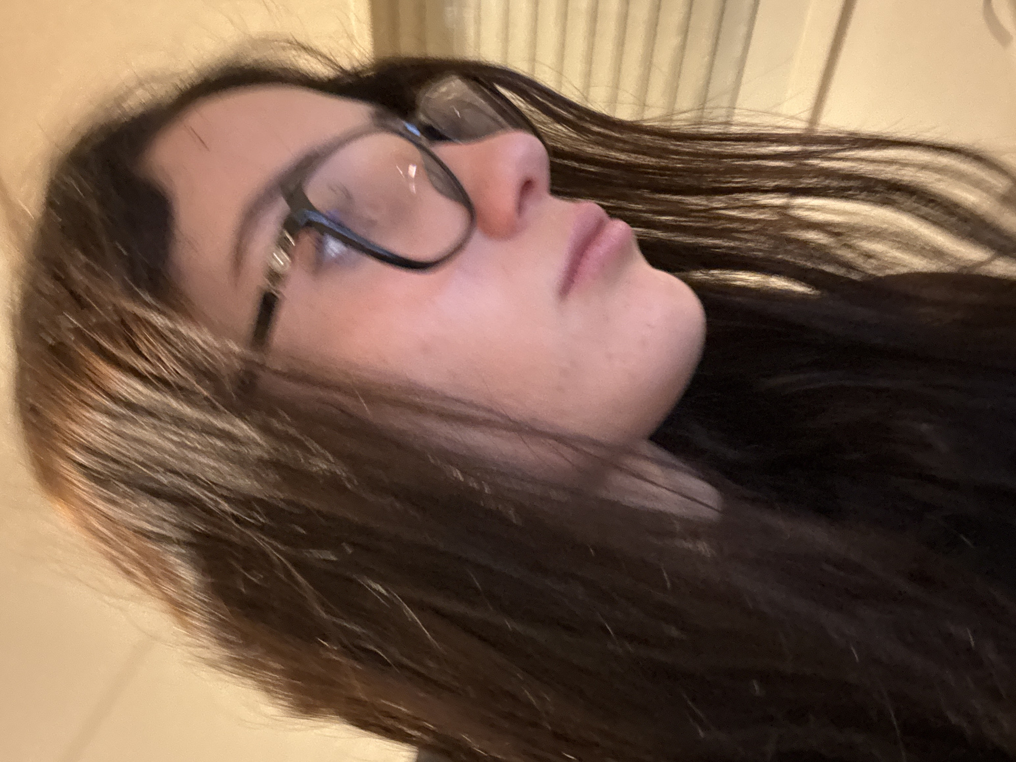 A close-up, slightly angled portrait shows a person with fair skin, dark hair, and wearing glasses. The person's face is in focus, showcasing their nose, lips, and the frame of the glasses. The hair is long and flowing, with some strands blurring due to motion. The background is out of focus, with hints of vertical lines suggesting a wall or blinds. The image does not reveal any location details. The person is not engaged in any observable actions.