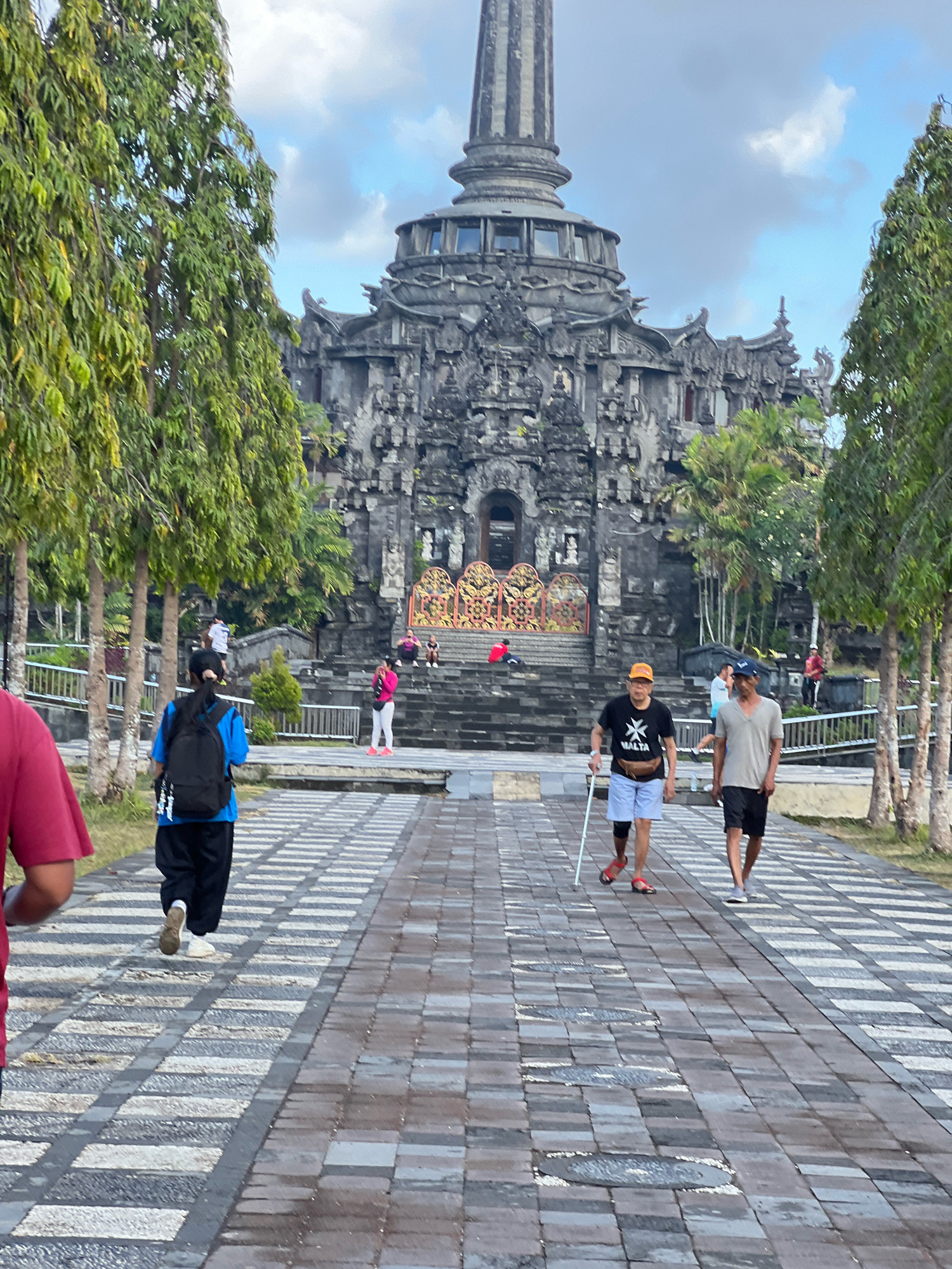 The image depicts a frontal view of a grand, stone temple complex in Denpasar, Indonesia, likely Bajra Sandhi Monument. In the foreground, a paved walkway with a patterned design stretches towards the temple. Two men are walking on the right side of the walkway. The man in the foreground on the right is wearing a grey t-shirt and black shorts, with a blue baseball cap. To his left, another man is walking with a white cane, wearing a black t-shirt with a white emblem and light blue shorts, and an orange baseball cap. A woman in a blue shirt and black pants with a black backpack is walking away from the camera on the left side of the walkway. Other individuals and trees are visible in the background, with the intricately carved, multi-tiered temple structure dominating the scene under a partly cloudy sky.