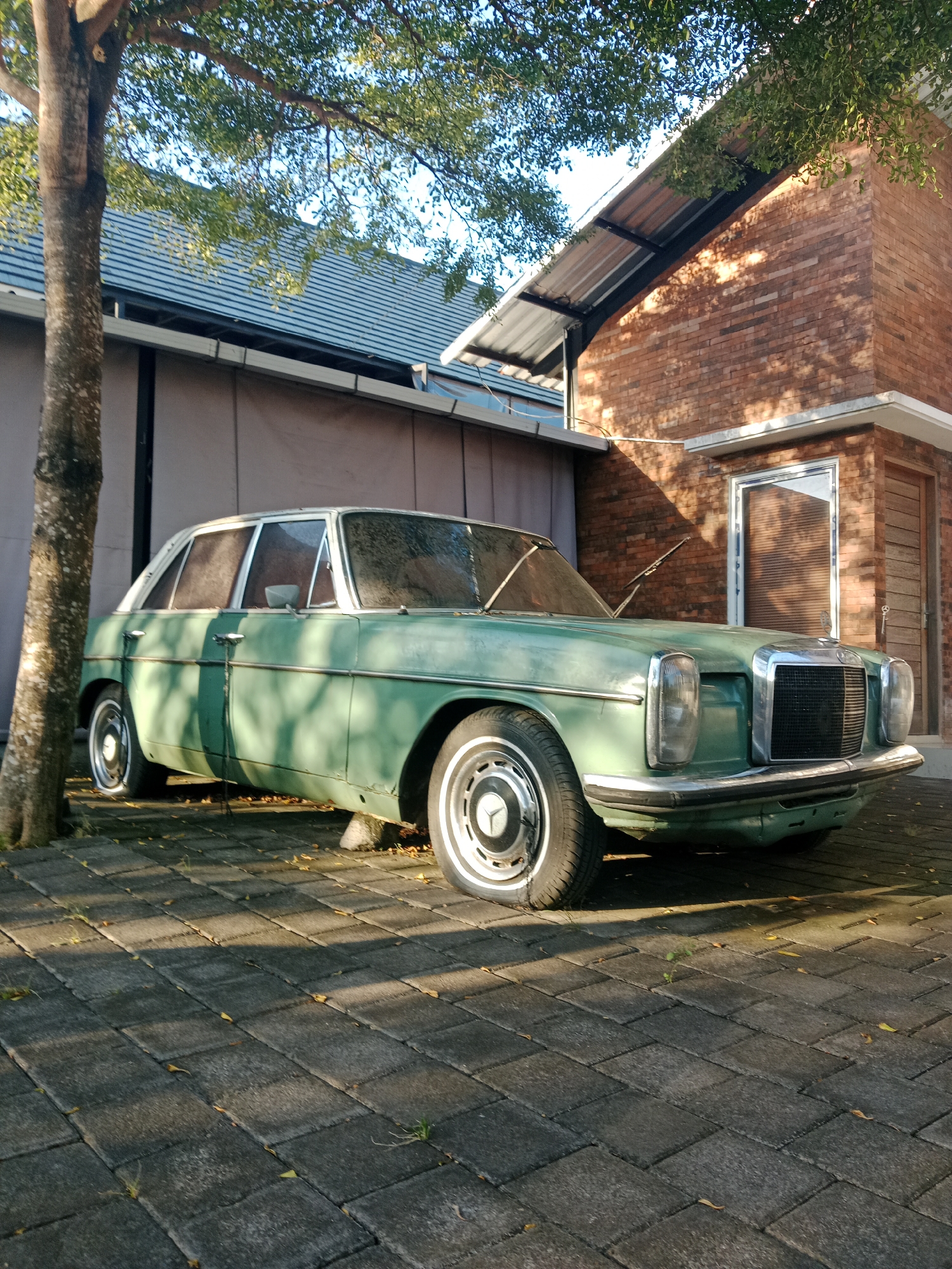 A vintage, light green Mercedes-Benz W115 sedan is parked on a brick-paved driveway in front of a building in Kuta Selatan, Indonesia. The car is positioned at an angle, facing the right side of the frame. Its chrome accents, including the grille and bumper, are prominent. The car's headlights are visible, and the windshield wipers are resting on the windshield. The building behind the car has a brick facade and a metal-roofed awning. A tree with green foliage is to the left of the car, casting shadows on the vehicle and the driveway. The driveway is composed of rectangular bricks, and scattered leaves are visible on the ground. The scene is bathed in natural light, suggesting it is daytime. No people are present in the image.