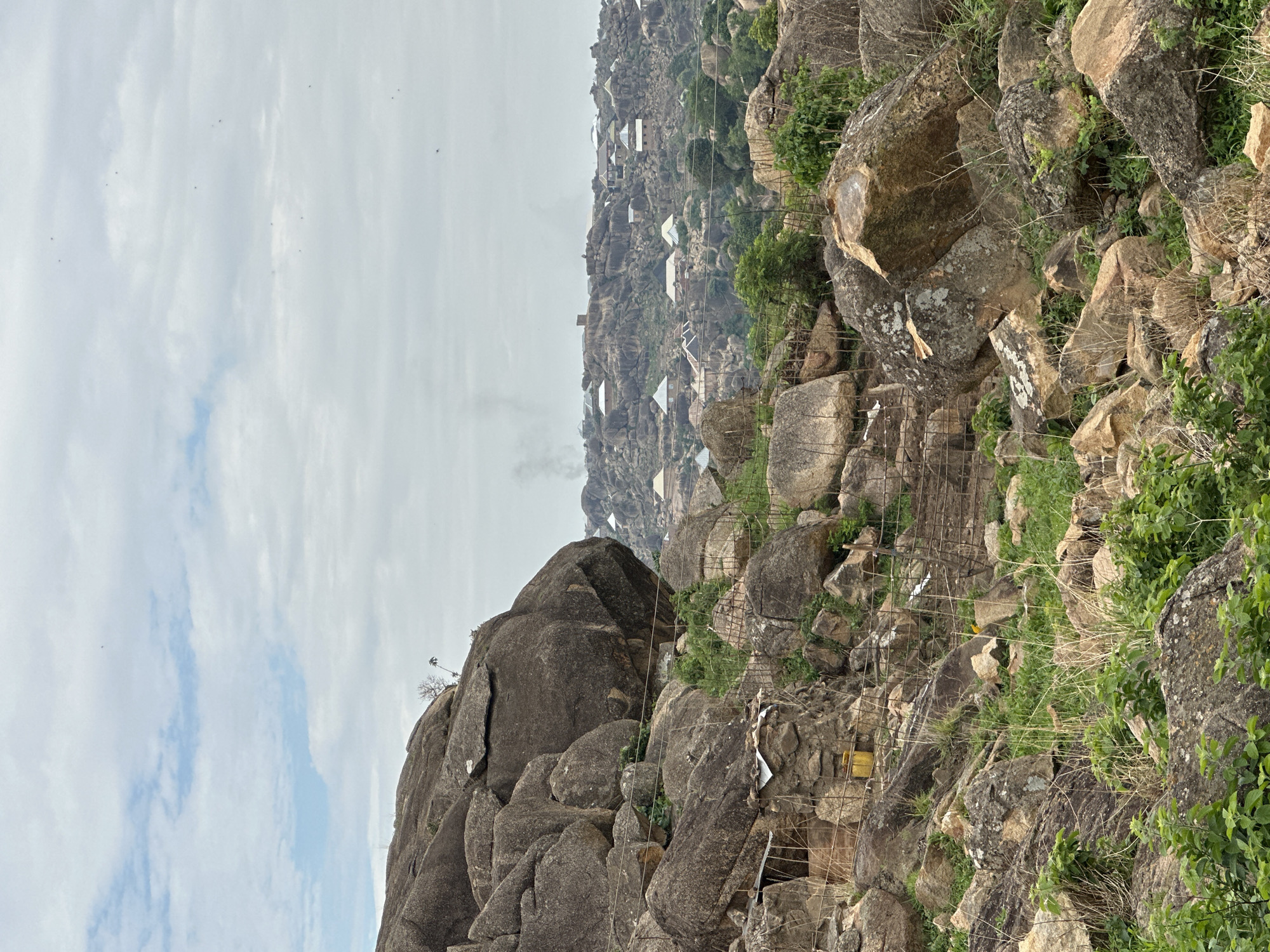 This image presents a wide view of a rocky, hilly landscape with human habitation, consistent with the geography of Jos, Nigeria. The sky occupies the upper portion, appearing largely overcast with patches of lighter blue clouds. Multiple small, dark specks, likely birds, are visible in the upper sky.
The foreground and midground are characterized by numerous large, weathered granite boulders and rocks varying in size, shape, and color, ranging from dark grey to reddish-brown, with some showing patches of white lichen. Green vegetation, comprising grasses and small bushes, grows sparsely between and over the rocks. A section of a wire fence is visible in the mid-left and extends towards the mid-right. In the lower-left, a small, rudimentary structure with a corrugated metal roof is built into the rocks, and a yellow container rests near another rock-built enclosure.
The background reveals an extensive village or settlement built directly into and among a dense field of similar rocky hills and inselbergs. Numerous small, single-story structures with light-colored, possibly corrugated metal, roofs are interspersed throughout the rocky terrain. Several plumes of light grey smoke are visible rising from the distant settlement, suggesting ongoing human activity such as cooking or burning. No individuals, specific actions, or direct interactions are discernible within the frame.