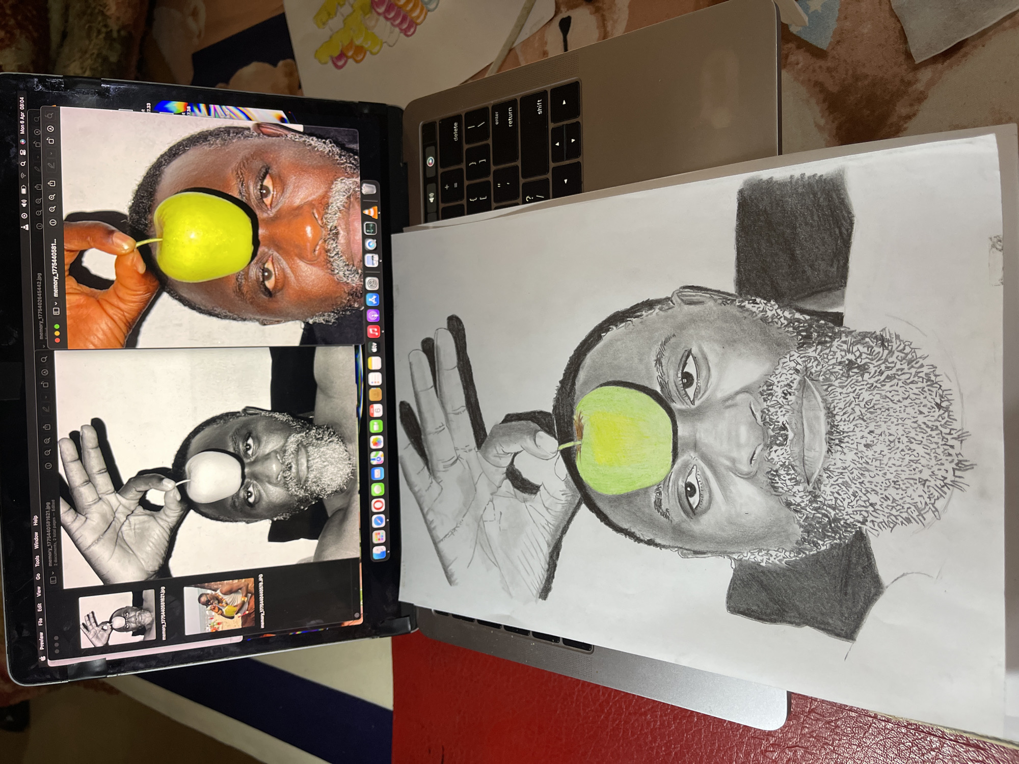 The scene depicts a drawing in progress, placed next to a laptop displaying the source image. The subject of both the photograph on the laptop and the drawing is a Black man holding a green apple in front of his eye. The man has a grey beard and is wearing a dark shirt. The drawing, executed in graphite, closely replicates the source photograph, though some details are simplified.
The laptop displays the photo being referenced, along with thumbnails of similar images. The laptop is resting on a red surface, which is possibly a desk or a table. The overall environment appears to be indoors. There is no location mentioned that specifies it being in Jos, Nigeria, or any other location.