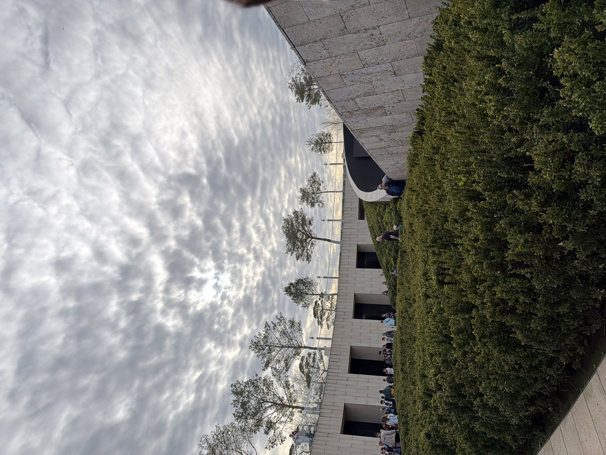 The image shows a view of the Park Krasnodar in Krasnodar, Russia. A long line of people extends alongside a tall, thick hedge next to a stone building with a series of doorways and windows. Trees are planted behind a low glass barrier. The sky is filled with streaky, overcast clouds.