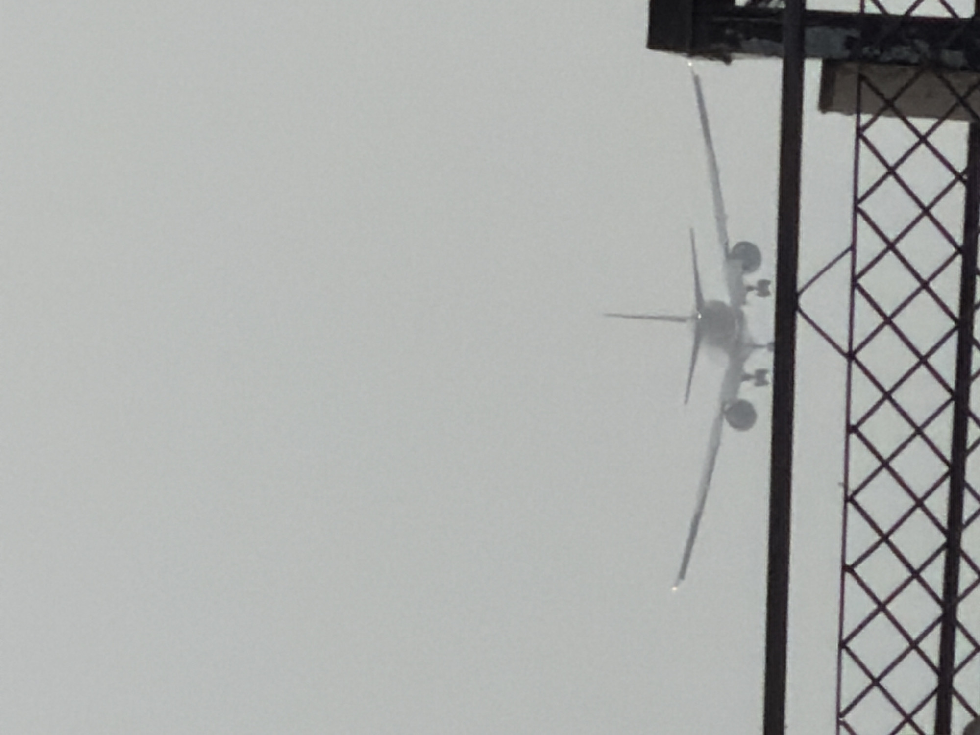 An airplane is seen from below, approaching for landing. The sky is overcast, and the view is partially obstructed by a wire fence and a dark railing. The scene appears to be within the vicinity of an airport. This perspective suggests the photograph was taken from a location near Indira Gandhi International Airport in Delhi, India, potentially from a public viewing area or a nearby overpass.