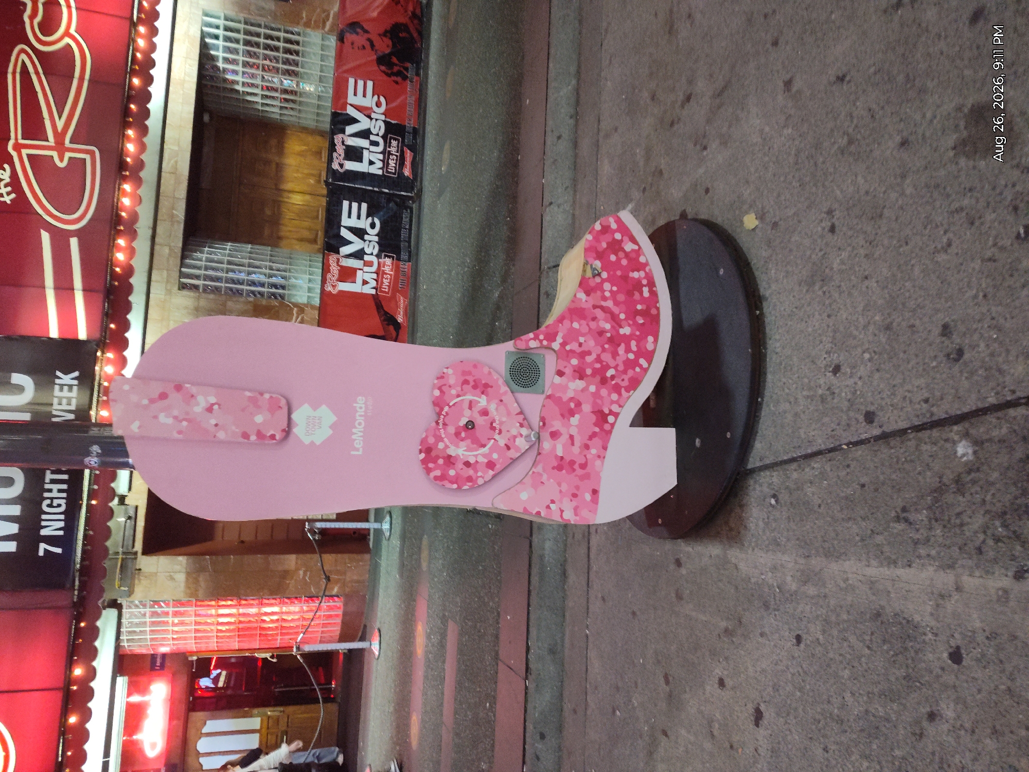 A tall, pink, boot-shaped sculpture with a heart-shaped button on its side stands on a sidewalk in front of a nightclub with a red awning. The sculpture has a pattern of pink and white confetti on it. The nightclub has signs advertising "7 Night Week" and "The Rock". There are people walking in the background, and banners advertising "Live Music" are hanging in front of the nightclub. The date stamp on the image indicates August 26, 2026, at 9:11 PM. The scene is likely in a busy entertainment district.