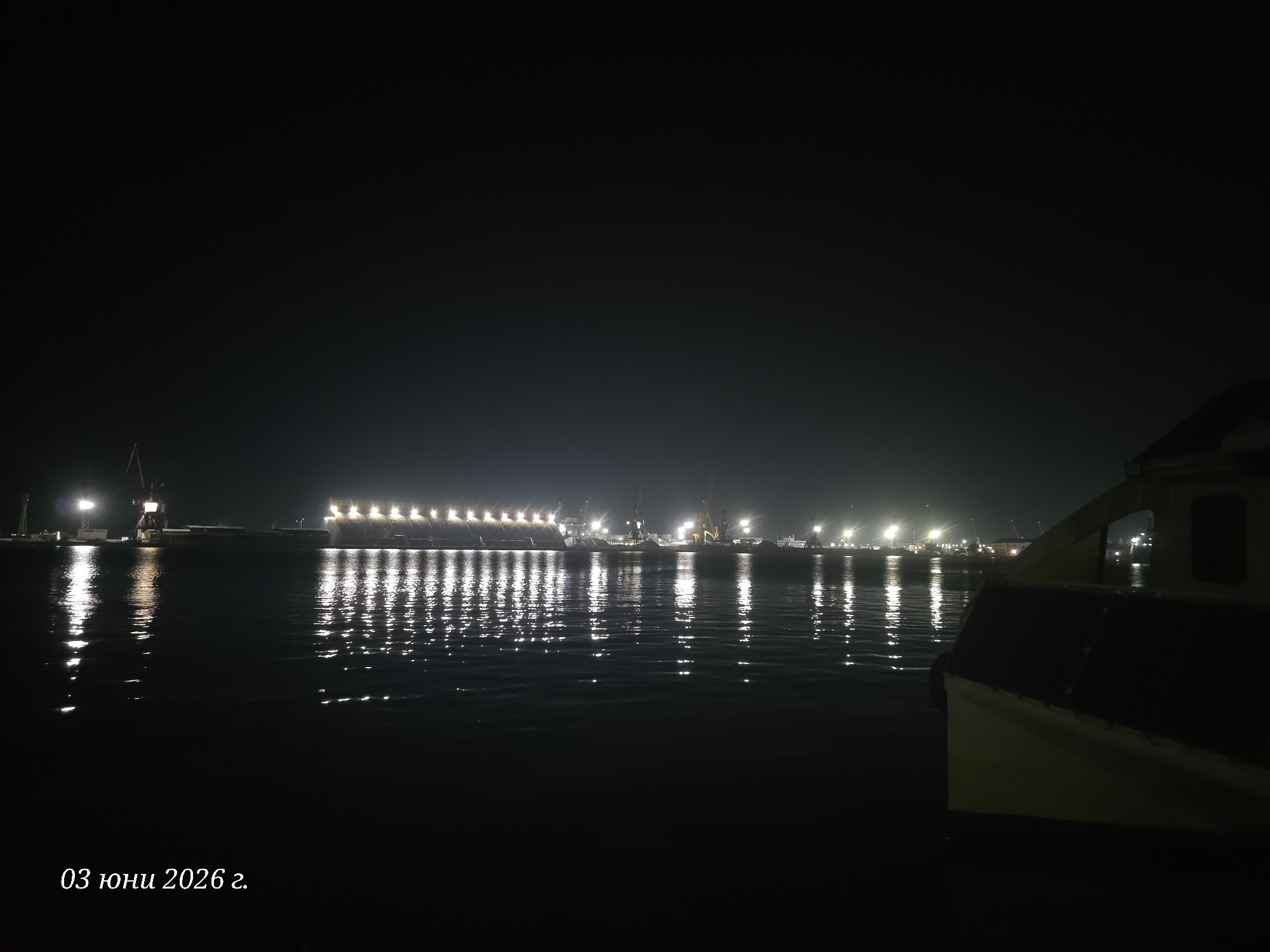 The image presents a serene nighttime view of a harbor or port, identified as Burgas, Bulgaria. The vast, dark sky dominates the upper portion, suggesting a clear night with no visible celestial bodies.
In the immediate foreground on the right side, a portion of a boat or vessel is visible, characterized by a light-colored, possibly yellow, hull section above a darker, shadowed lower part. The boat appears to be stationary, either docked or moored.
The main subject of the photograph is the expanse of dark water, which perfectly reflects the numerous artificial lights from the distant shoreline. These reflections create shimmering, elongated vertical streaks on the water's surface, dancing with the gentle ripples and contributing significantly to the visual depth and atmosphere of the scene.
Along the distant shore, a line of bright, white artificial lights illuminates the area. These lights appear to belong to a dock, industrial structures, or potentially large ships. Several cranes and other port infrastructure can be faintly seen silhouetted against the light. The arrangement of the lights suggests a bustling port area, even under the cover of night.
The overall scene conveys a sense of peaceful nocturnal activity in a maritime setting. In the bottom left corner of the image, white text clearly displays "03 юни 2026 г.", indicating the date "03 June 2026".