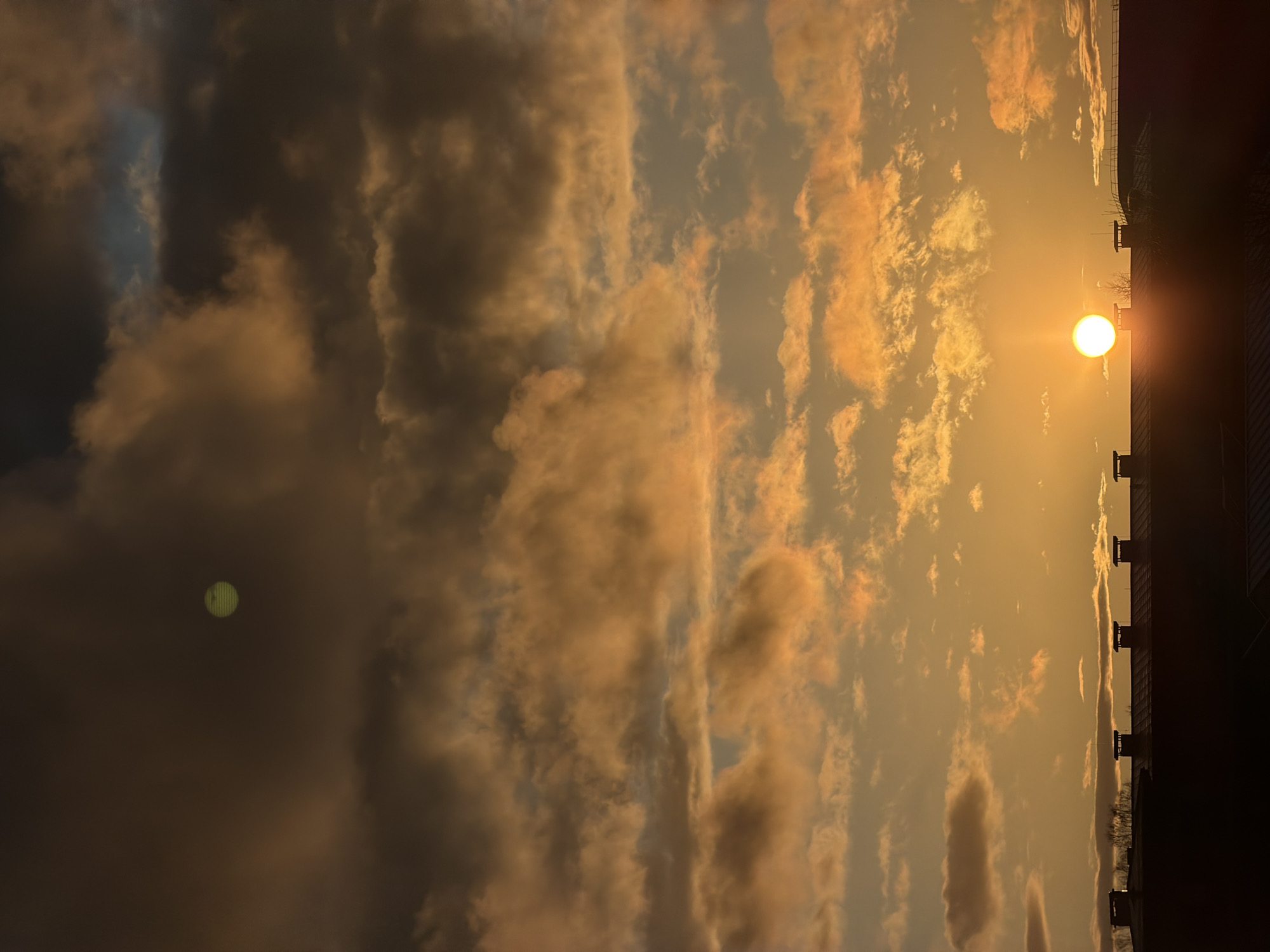 The image shows a vertical view of a sunset in Dreiliņi, Latvia. The sun is a bright orange sphere, positioned near the edge of a dark building on the right side. The sky is filled with clouds that are varying shades of orange and gray. A green, blurry orb is visible within a dark cloud on the left.