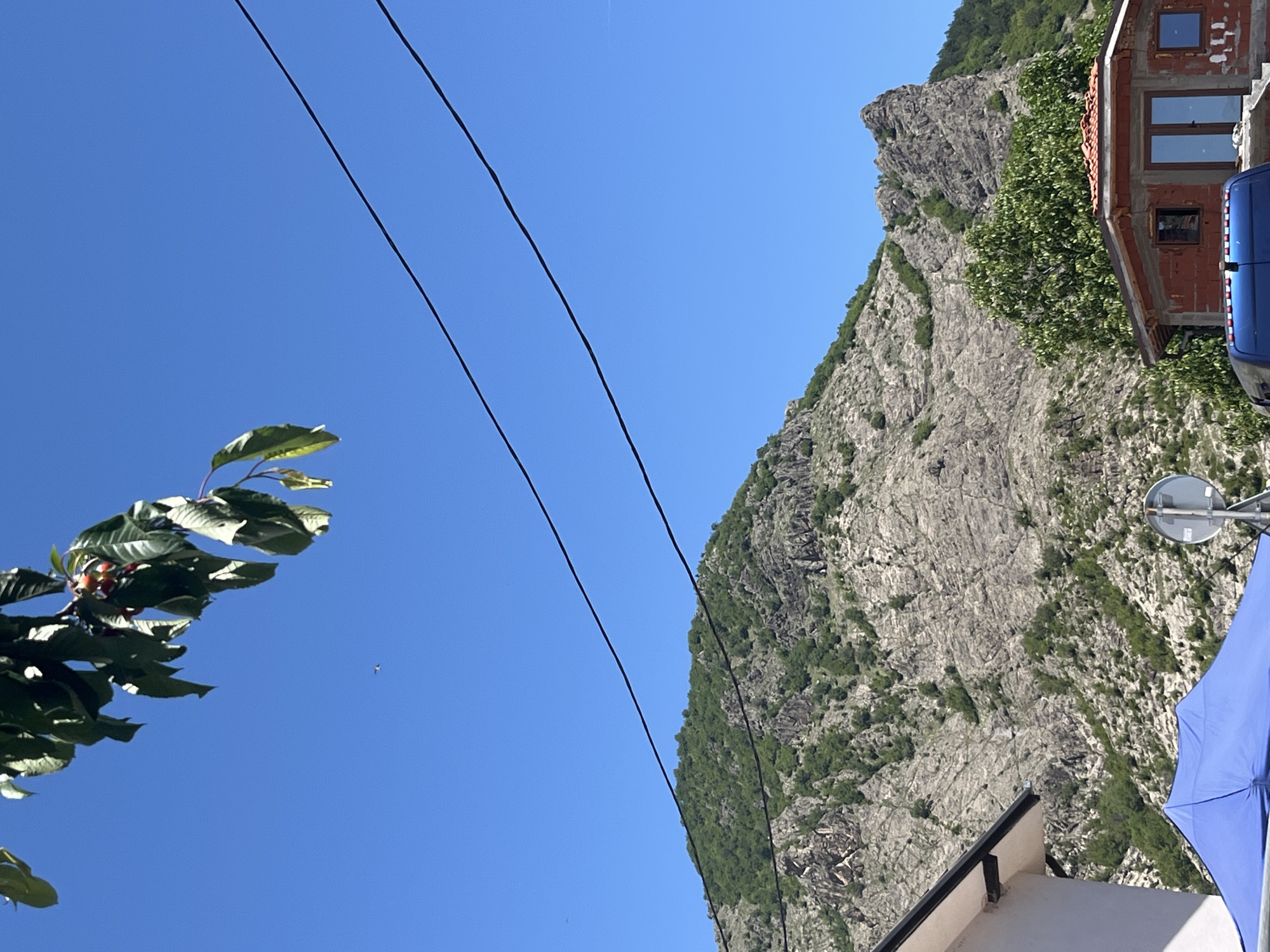 The image is an upward-angled view in Bukovo, Bulgaria, featuring a mountain slope covered with vegetation. A portion of a building with visible windows and a blue van is on the right side of the image. Three power lines span the top of the frame from left to right. The sky is clear and blue. Leaves and branches of a tree or bush are visible in the lower-left corner. Part of a white building with a black roof edge and a blue umbrella is visible in the lower right. A round mirror on a pole is located next to the umbrella.