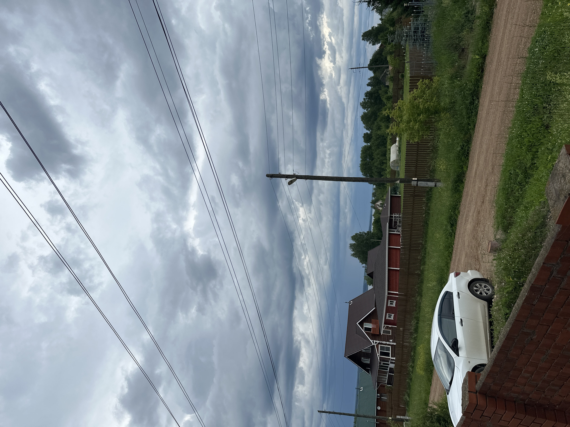 The image captures a scene in the city of Ilyinsky, Russia, under a heavily overcast sky. In the foreground, a white sedan is parked on a dirt road. Behind the car, a wooden fence encloses several rural houses, characterized by their dark-colored roofs and red or green siding. The background is dominated by a dense line of trees, with power lines crisscrossing the sky. A wooden utility pole with a light fixture is visible on the left side of the image. The overall atmosphere is subdued, likely due to the cloudy weather.