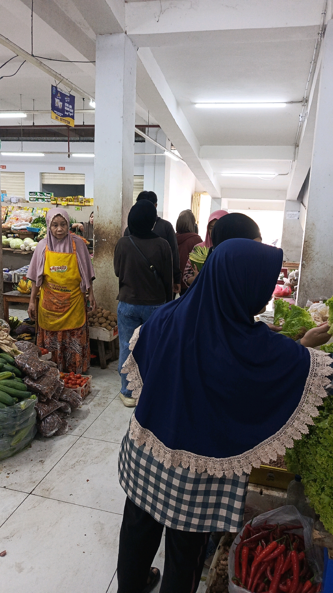 The image depicts a bustling indoor market scene in Nologaten, Indonesia, likely a wet market or a section of a larger market. In the foreground, a person wearing a dark blue hijab with a lace trim and a checkered apron is seen from the back, reaching for produce, possibly lettuce or other leafy greens. Behind them, a line of individuals, mostly women in hijabs of various colors, are queuing. To the left, a vendor, an elderly woman wearing a pink hijab and a yellow apron with "Masako" branding, stands attentively behind a stall piled with vegetables. Various produce, including cucumbers in plastic bags, tomatoes in baskets, and what appear to be dried goods, are visible on the floor and shelves. The market environment is characterized by concrete floors, white pillars, and fluorescent lighting. The overall impression is one of active commerce within a traditional Indonesian market setting.