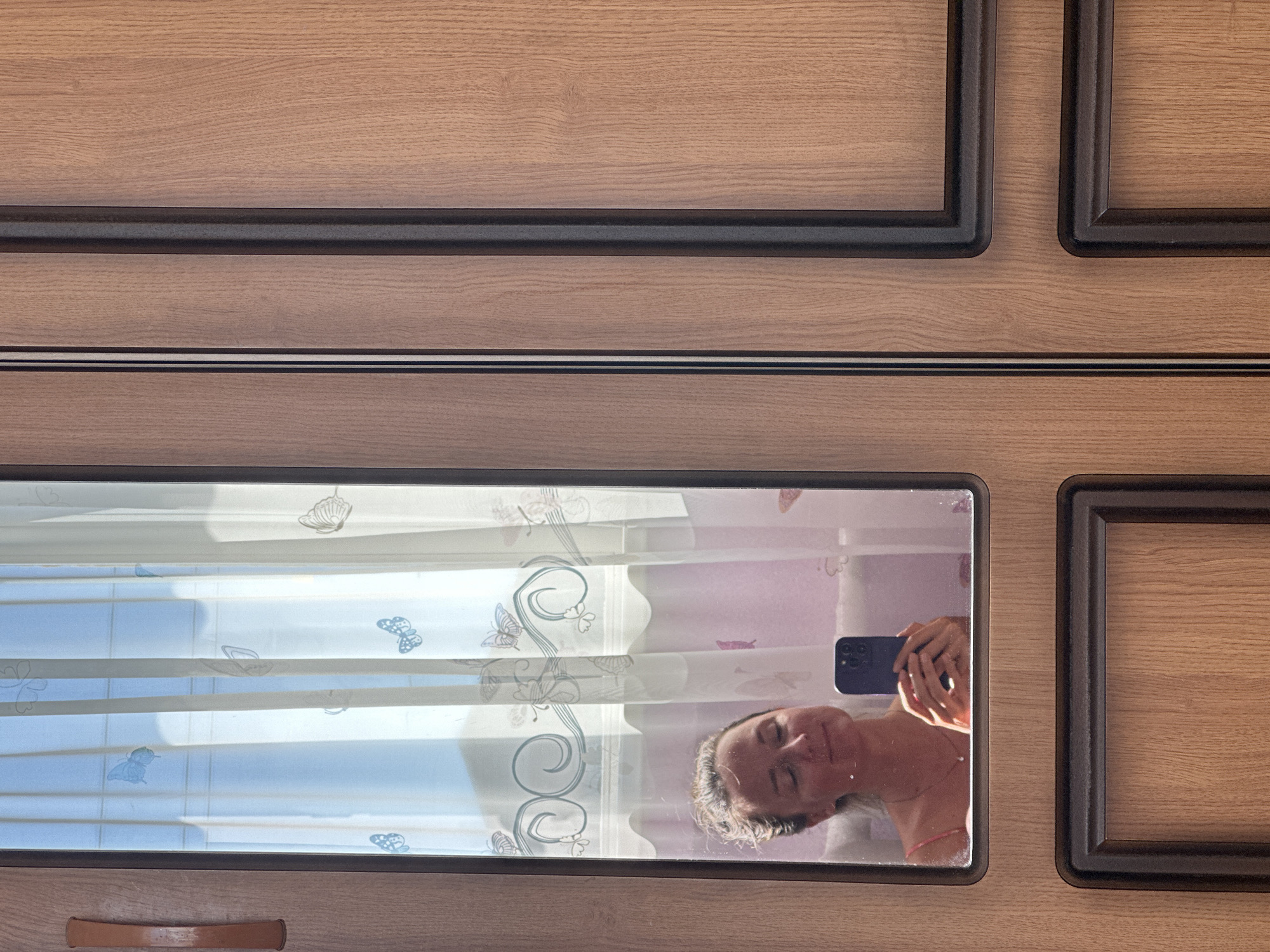 In the city of Rasskazovo, Russia, a woman is reflected in a mirror on a wooden wardrobe. She is holding a smartphone in her hands, seemingly taking a selfie, with her eyes closed and a slight smile on her face. She is wearing a necklace with a pendant and a pink strap visible from her clothing. Behind her, a sheer curtain with a butterfly pattern is illuminated by sunlight. The wardrobe itself has a wood-grain texture and is divided into panels.