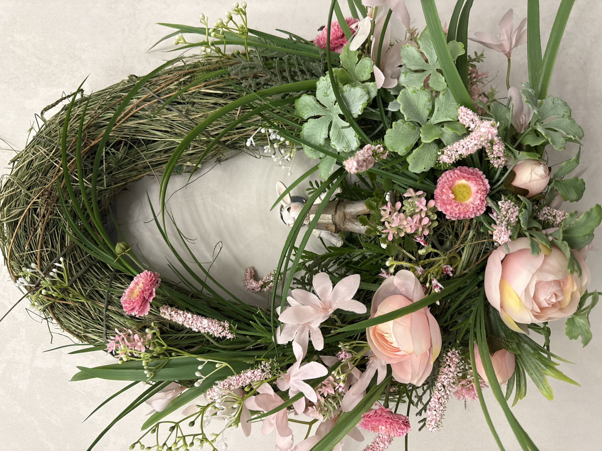 The image shows a floral wreath, predominantly composed of dried grass forming the circular base. Adorned on the right and lower left sections are arrangements of artificial pink flowers, including roses, daisies, and smaller blooms, interspersed with green foliage. A decorative figurine of a rabbit wearing a suit is partially visible within the wreath's interior. The background is a plain, light-colored surface.
There is no indication of location nor city in the image.