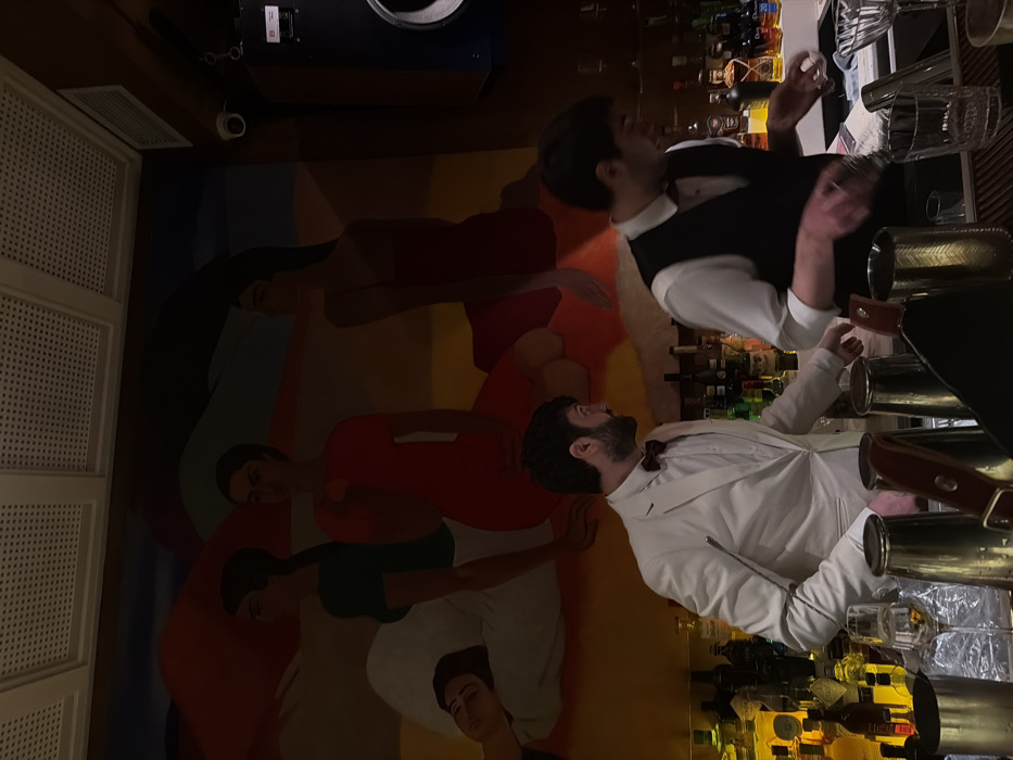 Two men are behind a bar, one in a white suit and bow tie, the other in a white shirt and black vest. Shelves of liquor bottles rise behind them. The floor features a colorful mural of figures in various shades of red, green, white, and blue. A patterned wall is partially visible on the left. The men appear to be working, possibly shaking or preparing drinks, with metal cocktail shakers in front of them.