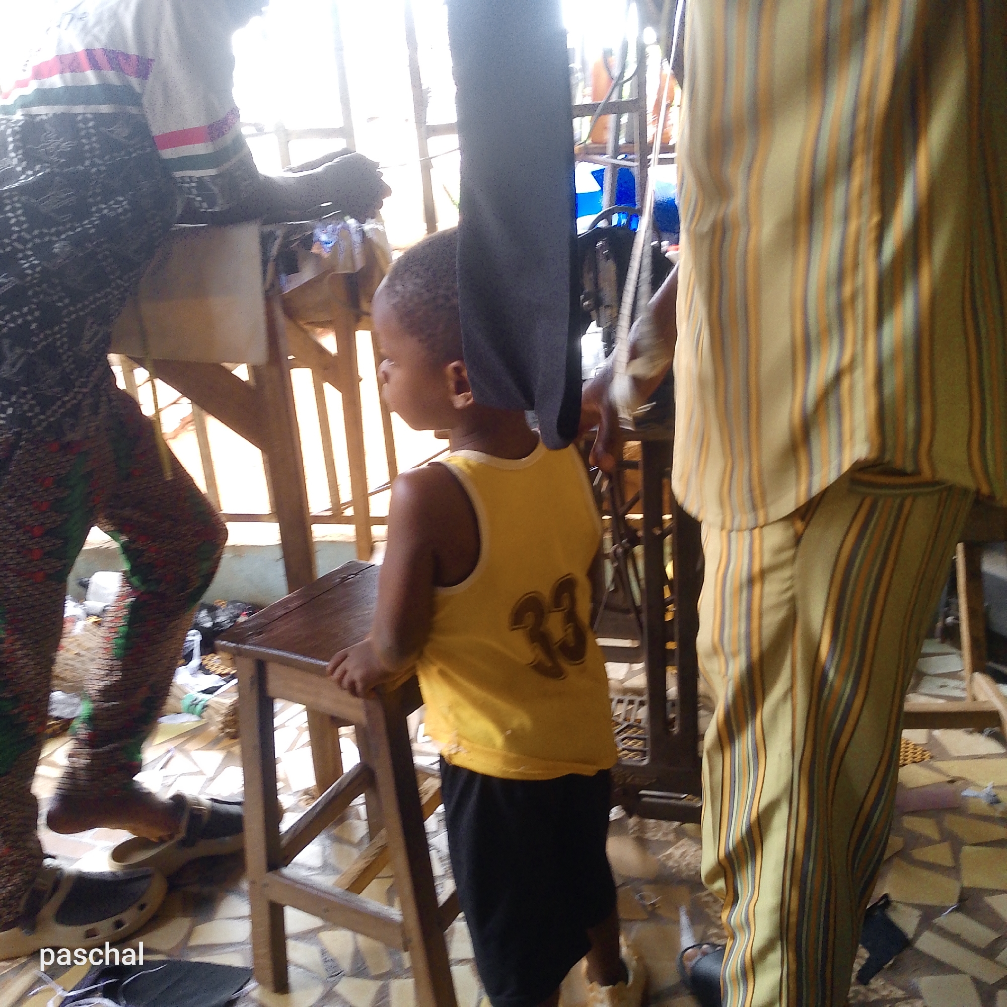 An eye-level interior shot depicts a child in a yellow tank top with the number "33" on the back, standing at a small wooden stool. A man to the left wears patterned trousers and a patterned top. To the right, another man is partially visible, wearing striped pants. A sewing machine is behind the child. The location is Unknown, Unknown. The floor is tiled with a triangular pattern. The name "paschal" is watermarked in the lower left corner.