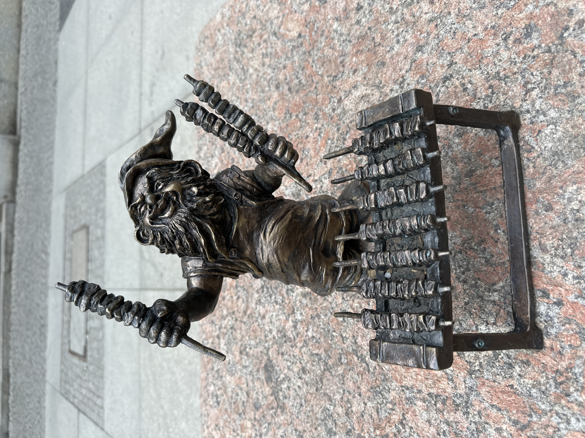 A bronze statue of a dwarf barbecue chef stands on a stone surface in Wrocław, Poland. The dwarf is depicted in mid-action, holding two skewers filled with meat in either hand, and stands to the side of a grill fully loaded with meat skewers. The dwarf wears a hat and has a thick beard. The grill is a rectangular, brick-like structure with a handle. The statue is weathered and shows signs of outdoor exposure. The stone surface around the statue is speckled with pink and grey tones. A section of a tiled exterior wall is visible in the upper left corner of the frame.