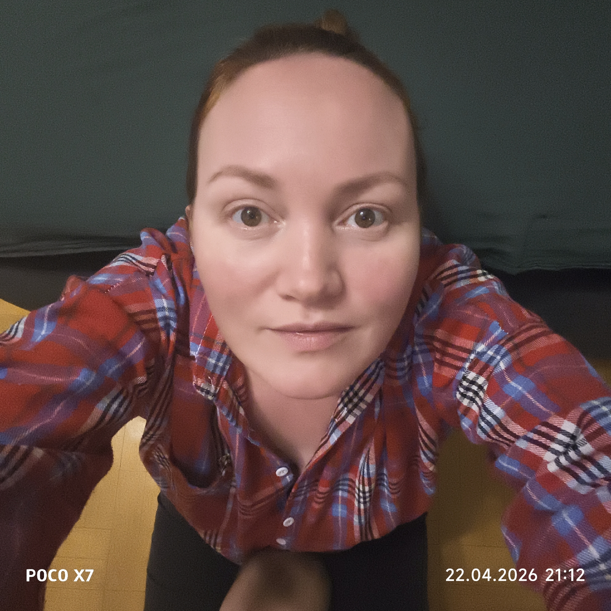 This indoor portrait features a person, likely a woman, looking directly into the camera from a low angle. She has light skin, reddish-brown hair tied back into a bun, and wide-set brown eyes with a neutral, calm expression. She is wearing a red, blue, and white plaid flannel shirt, with a few buttons undone at the neckline, over dark-colored shorts or pants. Her arms are visible, with her left arm extending towards the bottom left of the frame. She appears to be crouching or kneeling, with her head slightly tilted back, emphasizing the wide-angle perspective of the photograph.
The background behind her head is a dark green, possibly a sofa or wall, while the surface beneath her is a yellowish-brown floor. The wide-angle lens gives a slightly distorted view, particularly noticeable on her face.
Visible text overlays indicate "POCO X7" in the bottom left corner, identifying the camera device. In the bottom right corner, the date "22.04.2026" and time "21:12" are displayed, indicating the photo was taken in the evening. The setting is a domestic indoor space. The provided location context for this image is Chaikovsky, Russia.