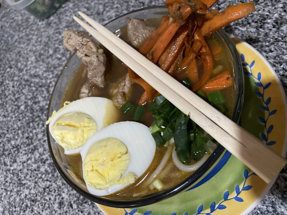 A bowl of ramen sits on a colorful, patterned plate. The ramen contains hard-boiled eggs, sliced scallions, and chunks of meat. Cooked carrots are piled on top. A pair of wooden chopsticks rests across the bowl. The countertop has a speckled gray pattern. A container holding some food is in the upper left corner.