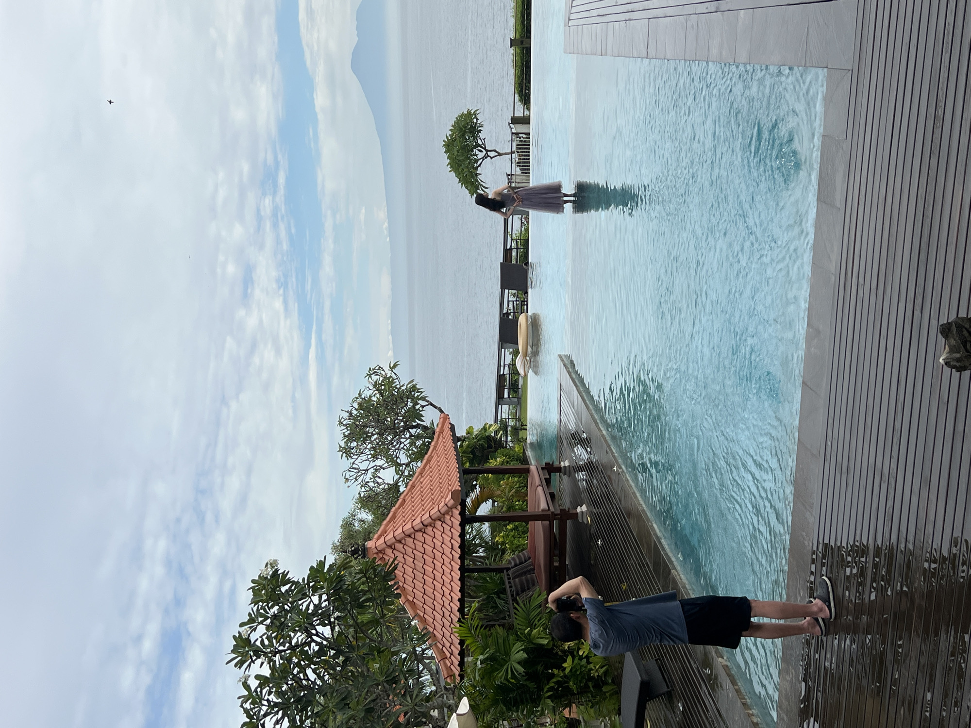 An outdoor scene captures a picturesque resort setting during daytime, likely in Kuta Selatan, Indonesia. The image is dominated by an infinity pool with clear blue water, which merges seamlessly with a vast expanse of ocean or sea in the background. Distant mountains or large landmasses are visible on the horizon.
Two individuals are present. A person, appearing to be a woman, stands gracefully in the shallow end of the pool, facing away from the viewer and looking out towards the panoramic ocean view. She is wearing a flowing, light-colored dress or skirt. On the wooden deck bordering the pool, another person, likely a man, is positioned with his back to the camera, holding a black digital camera. He appears to be photographing the person in the pool, suggesting a photo shoot or a leisure activity capturing vacation memories.
The pool deck is made of dark wooden planks. To the lower left of the frame, a traditional-style gazebo with an orange-red tiled roof and wooden pillars is nestled amidst lush green tropical foliage. Various trees and plants frame the scene, including a distinctive small, rounded tree near the pool's edge in the midground. The sky above is bright and clear, with scattered white clouds. A small bird is visible flying in the upper left portion of the sky. The overall atmosphere conveys tranquility and leisure in a beautiful tropical environment. No text is visible in the image.