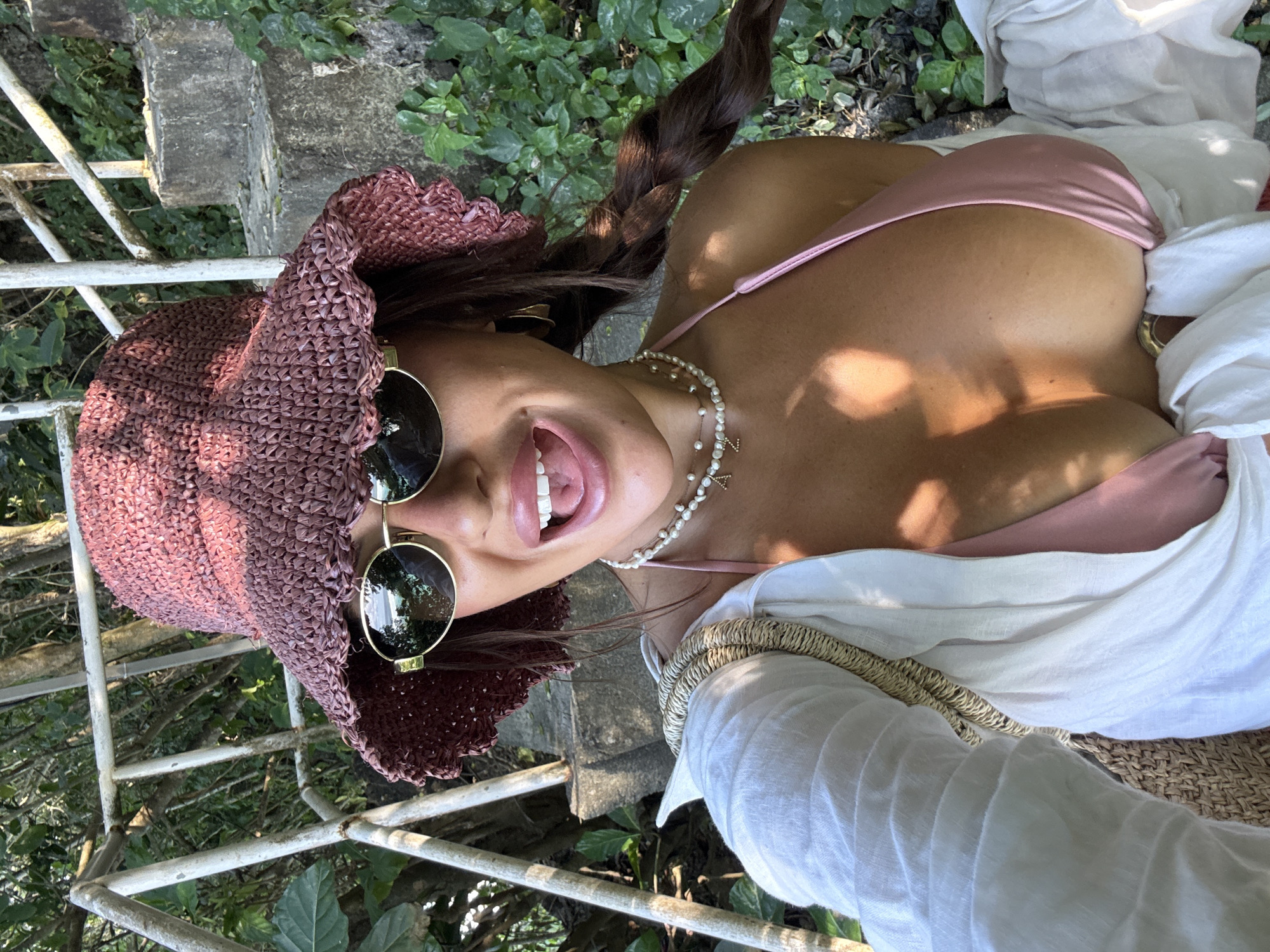 A woman is lying on a woven mat on stone steps in Kuta Selatan, Indonesia. She is wearing a pink bikini top, a white linen shirt with a woven bag strap, and a burgundy woven hat with round sunglasses perched on the brim. She also wears a pearl necklace. Her hair is styled in a braid. The background includes a white metal railing and lush green foliage.