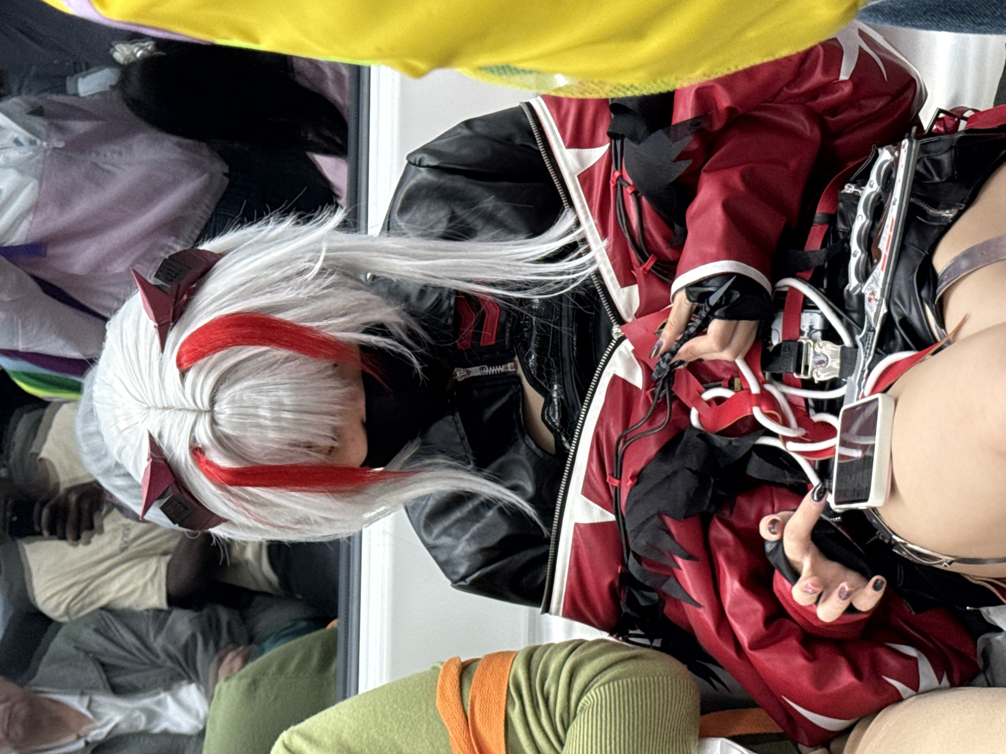 A person is wearing a white wig with red highlights, a black jacket with red and white accents, and holding a smartphone. The setting appears to be indoors, possibly at a convention or gathering. People and various colors of fabric are visible in the background, partially reflected in what appears to be a window or reflective surface.