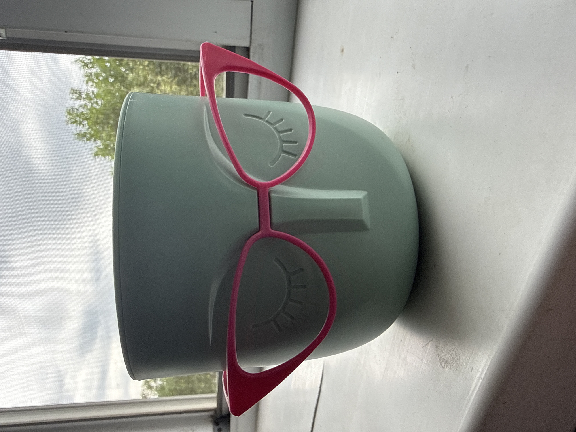 A close-up, eye-level shot shows a light green, ceramic pot shaped like a human head with a nose and closed eyes etched into its surface. The pot is wearing a pair of bright pink cat-eye glasses that are resting on the bridge of the nose. The pot is sitting on a white windowsill against a background of a window with a screen. Through the screen, a cloudy sky and green trees are visible. The image appears to have been taken indoors, possibly in a home or office, with natural light coming from the window. The location in the city of Tekeli, Kazakhstan is not identifiable from the image.