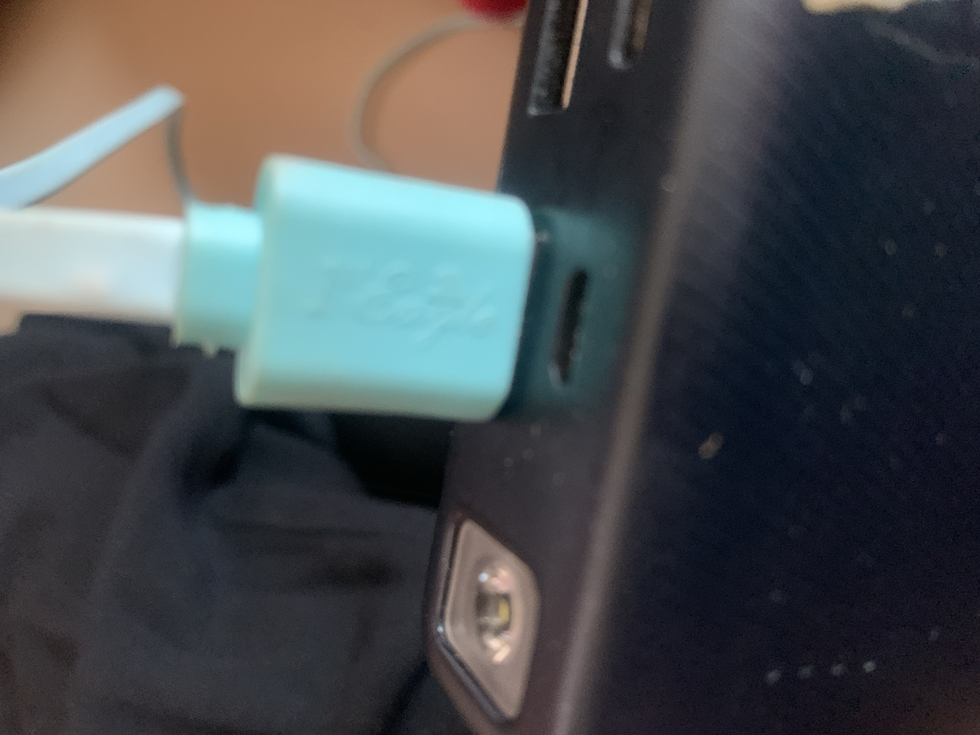 A close-up, slightly blurry, overhead shot shows a teal colored, rectangular object with a white tube emerging from the top and a light blue nozzle at an angle. The teal object has an embossed, unreadable logo on its surface. This object is resting on a dark, matte surface with several horizontal slots. To the left of the teal object, a small, rectangular, silver-colored light is visible, emitting a soft yellow glow. The background consists of dark, textured fabric on the left and a blurred, warm-toned surface on the right. There are no discernible people or interactions. The scene does not provide any specific indicators of its location in Jauro Yinu, Nigeria.