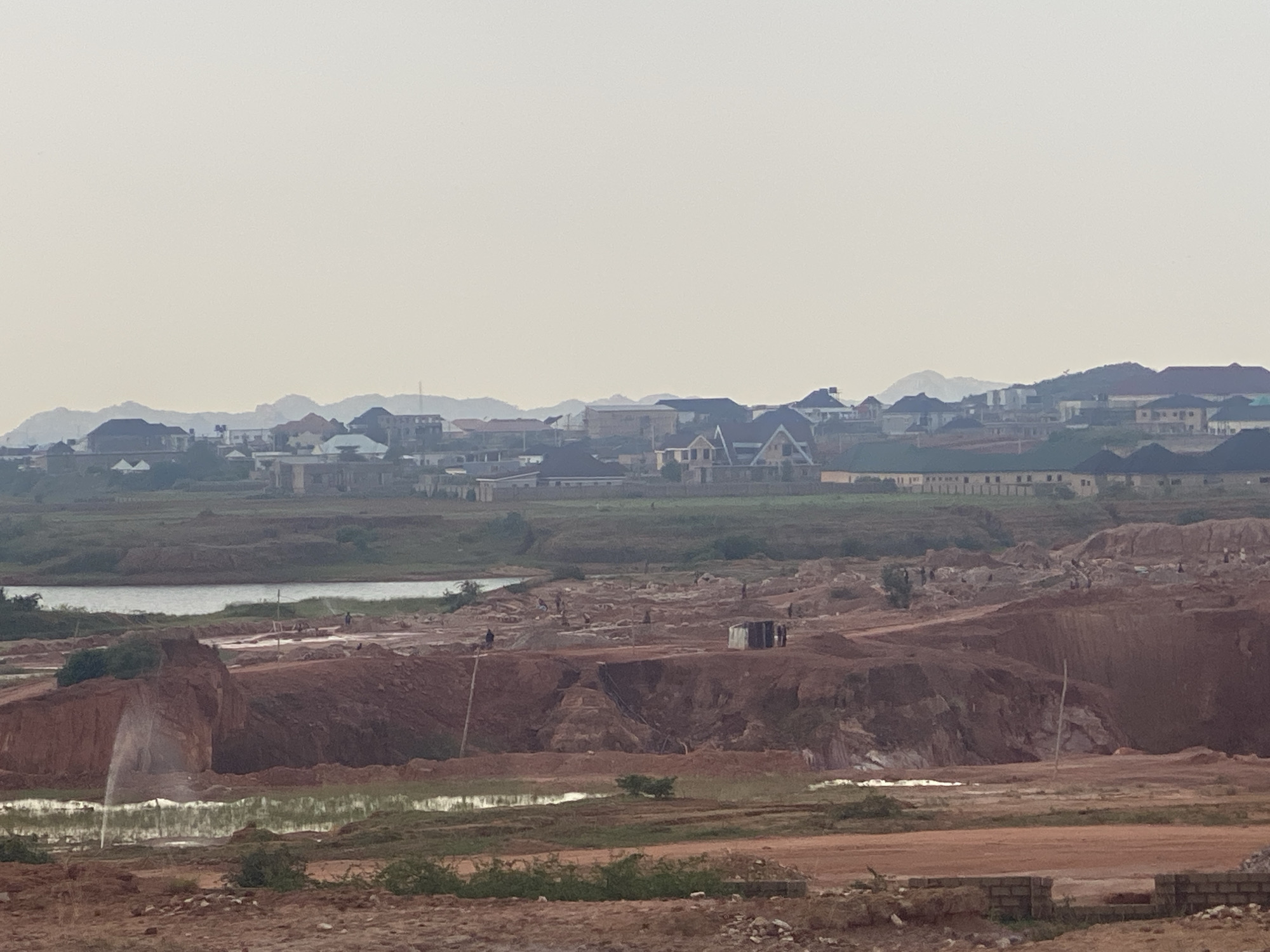 The image depicts a panoramic view of a construction site or quarry operation in Jos, Nigeria, characterized by large mounds of reddish-brown earth and several bodies of water, possibly retaining ponds. In the foreground, the ground is covered in loose soil and scattered building materials, including what appears to be a brick wall under construction. Mid-ground is dominated by the excavation site, with steep, eroded earth banks and intermittent sprinklers wetting the ground. Several individuals, appearing as small figures from this distance, are visible working within the site. A small, dilapidated structure, possibly a shelter or storage shed, is situated on one of the higher mounds. In the background, a lake or large body of water is visible, with more figures working near its edge. Further in the distance, a developing residential area with modern houses and buildings is visible, nestled against a backdrop of rolling hills and mountains, characteristic of the Jos Plateau. The sky is overcast, creating a diffused light that softens the landscape.