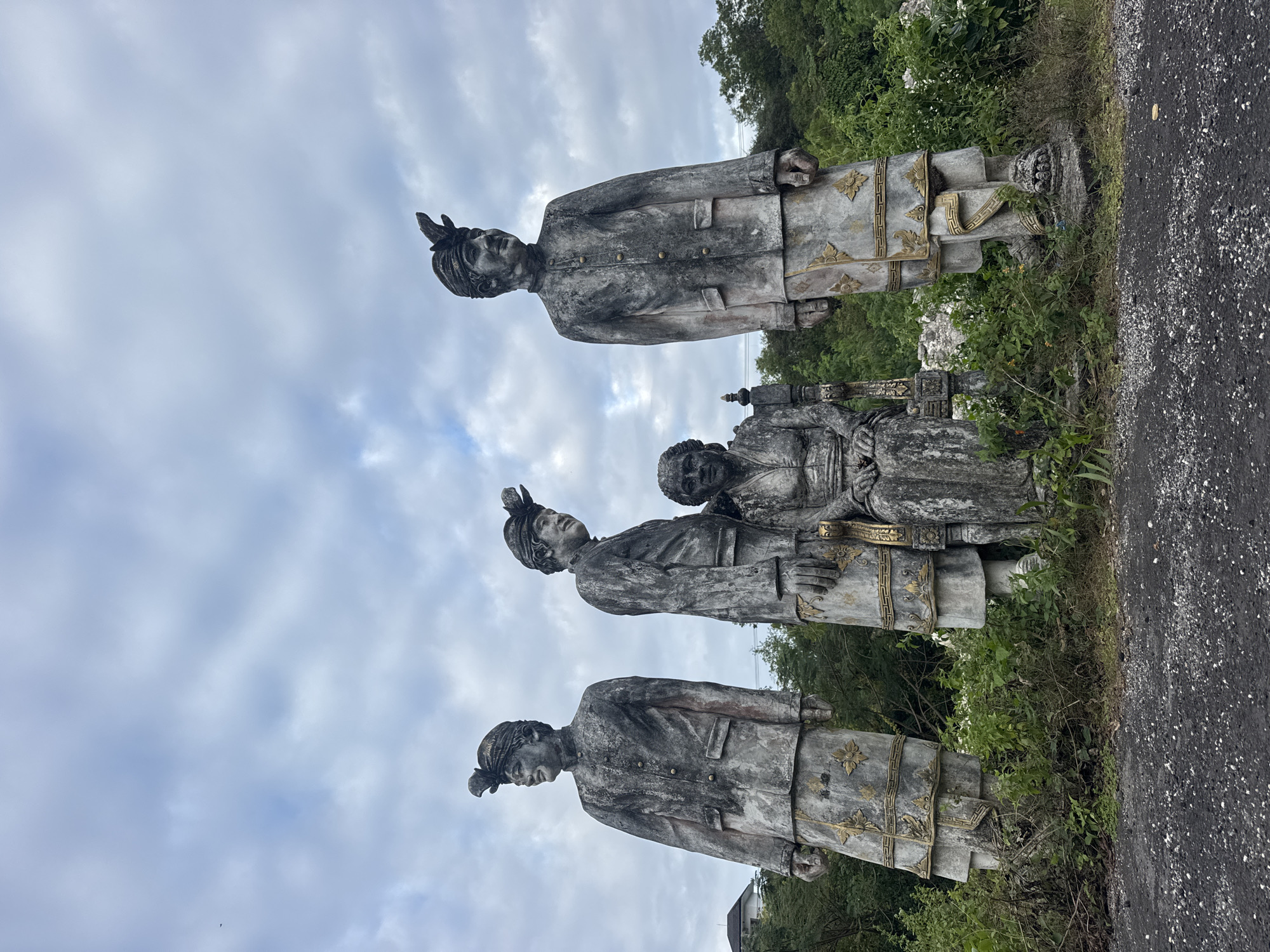 In Kuta Selatan, Indonesia, four weathered stone statues stand amidst overgrown vegetation and a cloudy sky. Three statues, depicting men in traditional attire with ornate headwear and jackets over patterned lower garments, are positioned to the left and right. The third statue from the left stands slightly behind the other two, and the fourth statue is to the far right. Between the second and third statues, a seated female figure is depicted, also in traditional clothing. The statues show signs of age and wear, with moss and dirt accumulated on their surfaces. The ground in front of the statues is rough and dark, likely asphalt or gravel, with scattered small white pebbles. Lush green foliage surrounds the base of the statues, with some branches and leaves reaching up to them. In the background, hints of buildings and dense green trees are visible beneath the overcast sky.