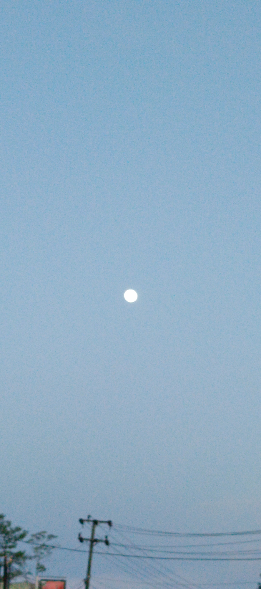 A vertical, slightly grainy, medium-shot photograph captures a pale blue sky with a small, bright white circle, presumably the moon, situated slightly above the center. In the lower portion of the image, a dark telephone pole with multiple wires running across it is visible. To the left of the pole, the blurry green tops of trees and a hint of a red structure, possibly a building or sign, are present. The scene appears to be an outdoor environment in Sukamulia, Indonesia.