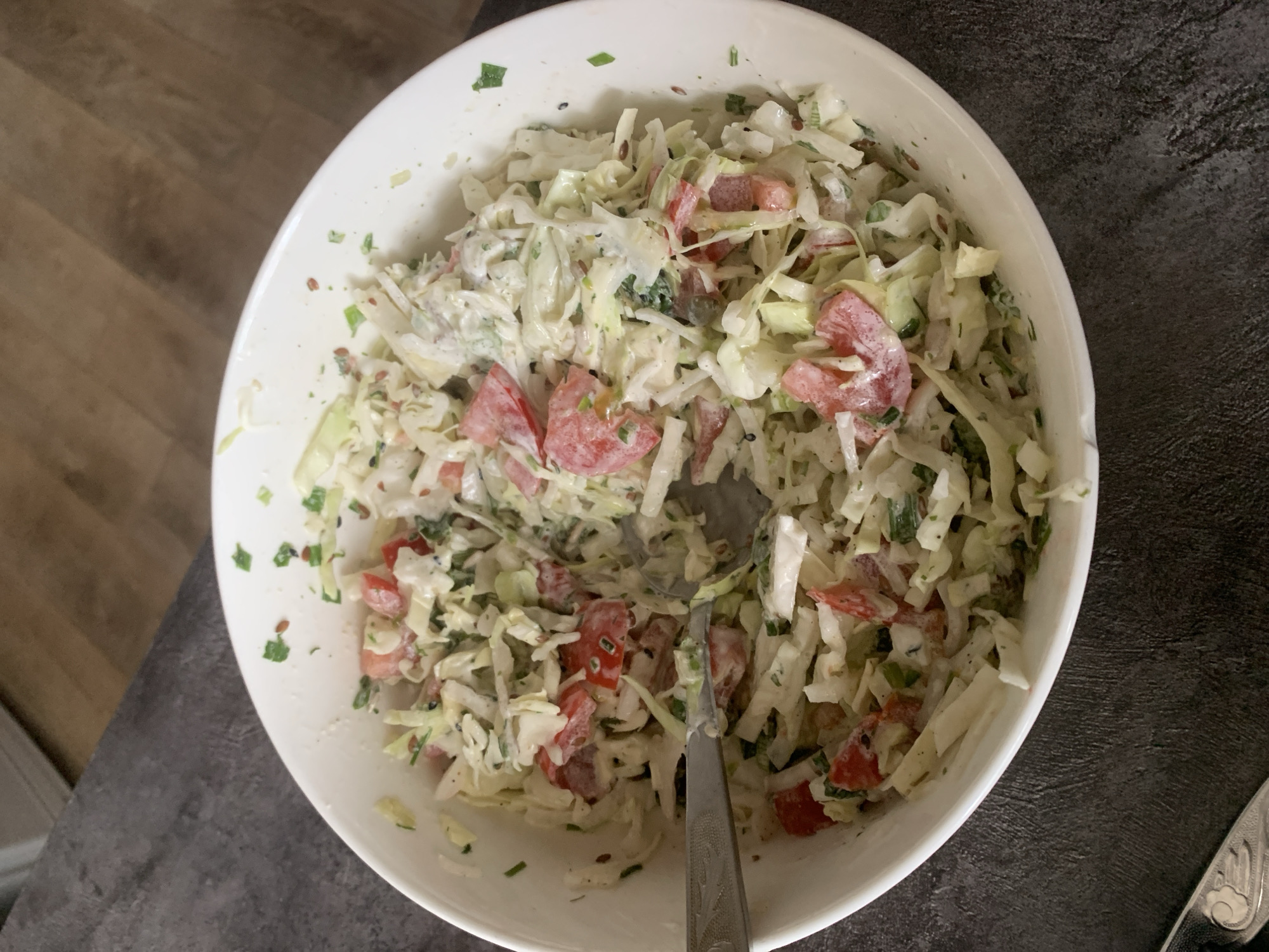 A close-up overhead shot shows a white bowl filled with coleslaw. The coleslaw is composed of shredded cabbage, diced tomatoes, and chopped green herbs, all coated in a creamy dressing. A silver fork rests on the edge of the bowl, partially submerged in the salad, suggesting it is being served or eaten. The bowl sits on a dark, textured surface. The background is out of focus, showing a hint of a wooden floor and a white object, possibly a cabinet or counter. There are no people or animals present. The scene is indoors, with natural lighting. The image does not provide any information that can be used to determine its location in Starodub, Russia.