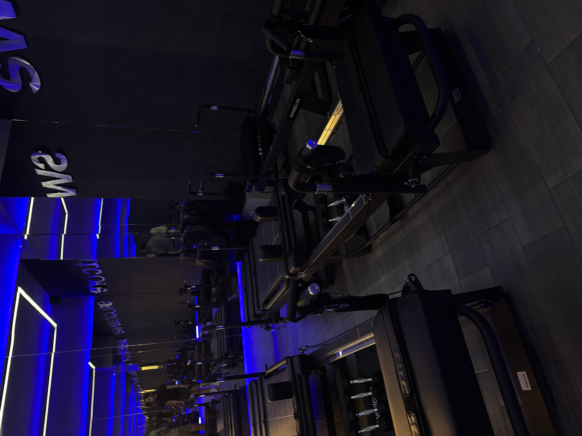 The image captures a gym interior in Puebla, Mexico. Rows of Pilates reformer machines, labeled "Sweatcore," are aligned along a mirrored wall. Blue LED lights illuminate the mirrors and create a neon effect. Reflections in the mirrors show individuals using the equipment. The floor is tiled with gray planks.