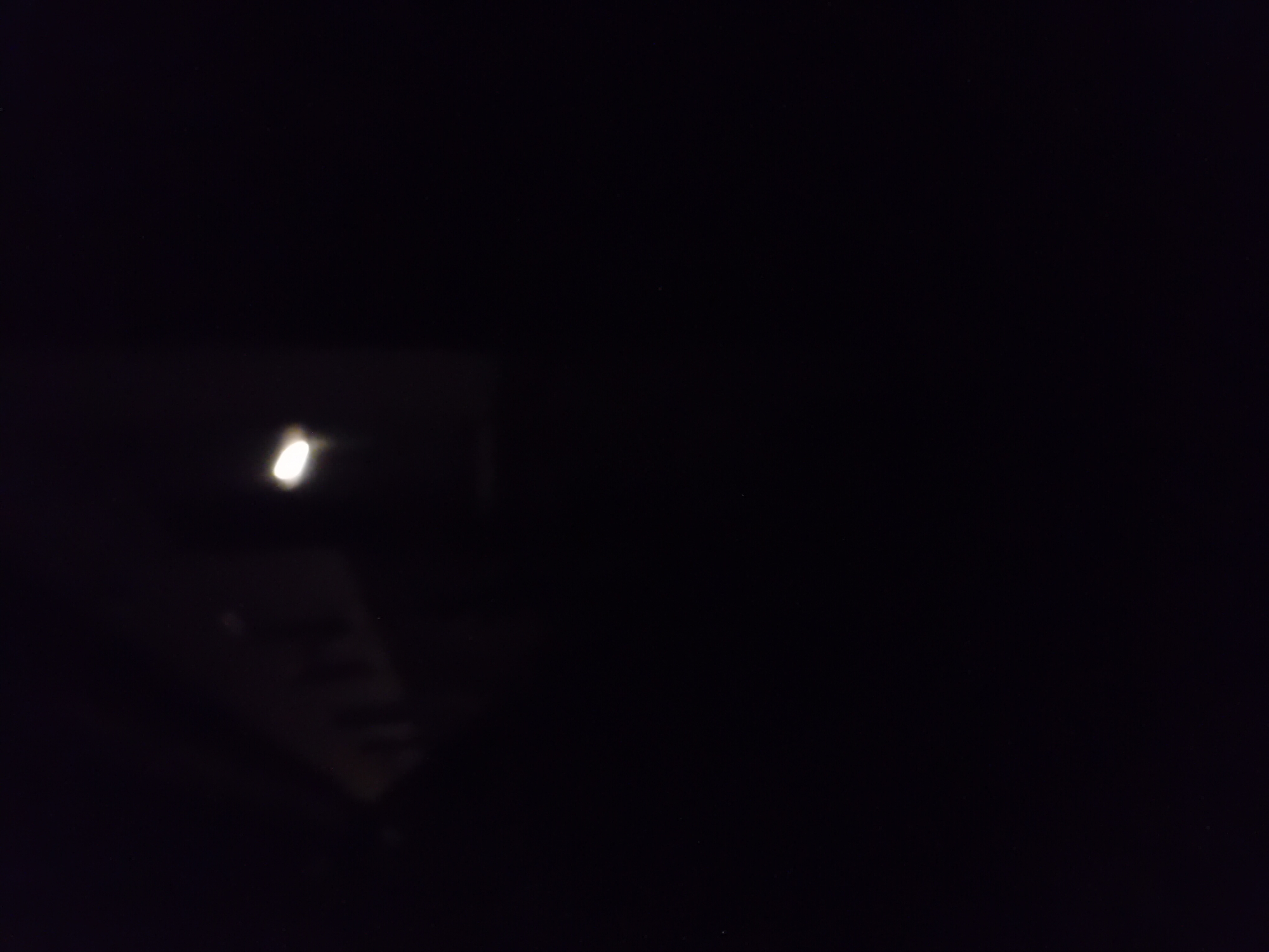 The image shows a very dark scene with a bright light source on the right. In the upper left portion of the image, a faint structure resembling a building is visible. The structure appears to have multiple rectangular shapes that could be windows or openings. The overall impression is that the photograph was taken at night, and the dark surroundings are a result of low light conditions. The light source on the right is out of focus and appears to be an artificial light.