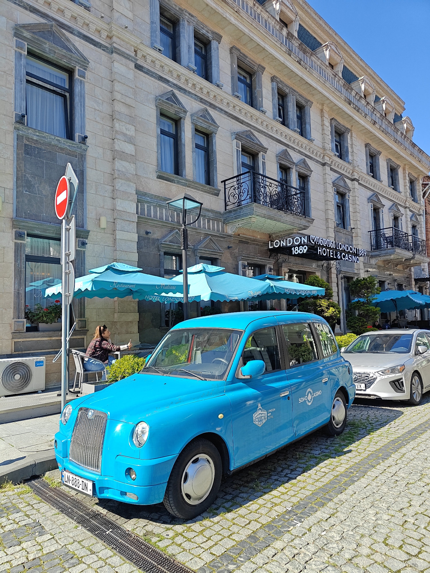 The image shows a bright blue London-style taxi parked on a cobblestone street in front of the London 1889 Hotel & Casino in Batumi, Georgia. The hotel is a multi-story building with light-colored stone facade, dark-framed windows, and iron balconies. A no-entry traffic sign stands next to the building. A woman is seated under a turquoise umbrella, apparently using a phone. A white Hyundai car is parked behind the taxi. The sky is clear.