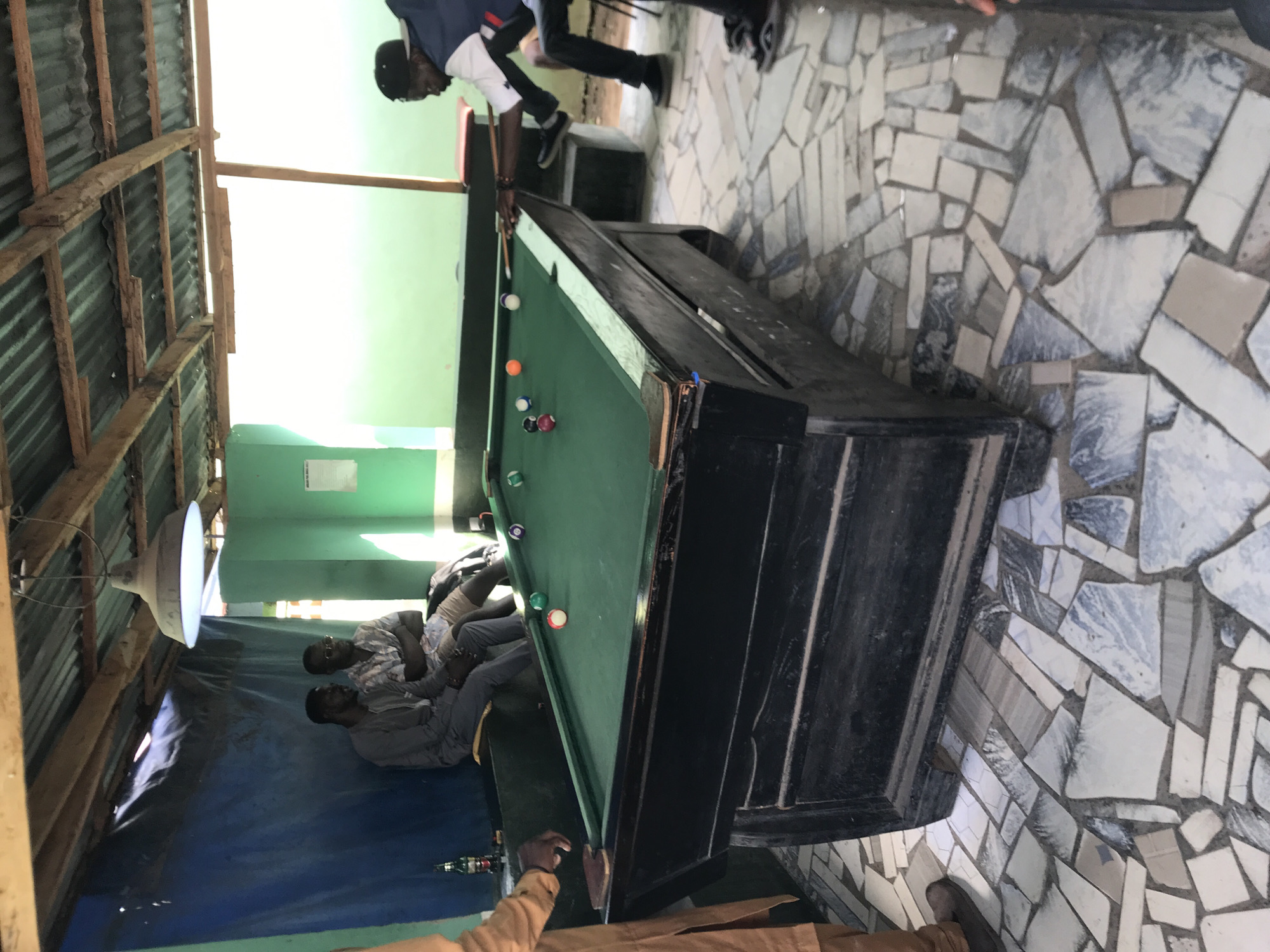 This image captures a casual indoor-outdoor setting in New Stadium, Nigeria, featuring a billiards game in progress.
In the foreground and midground, a dark-colored pool table with a green felt surface occupies the central space. Several colored pool balls are scattered across the table, including red, green, orange, and purple. A man, wearing a dark baseball cap and a white and navy blue jacket, is positioned on the right side of the table, holding a wooden pool cue as if preparing to strike a ball. His legs are visible, clad in dark trousers and black shoes. To the far left, a person partially visible, wearing a light brown or tan tunic, extends their right arm towards the table.
In the background on the left, three men are seated on a dark-colored bench or ledge. The man furthest left wears a grey long-sleeved garment, while the man next to him wears a patterned short-sleeved shirt and glasses. The third man is less distinctly visible. A dark bottle, possibly a beverage, rests on a ledge near the seated men. The wall behind them is partly covered by a dark blue tarp or fabric, transitioning to a light green painted wall further right. A single, inverted cone-shaped pendant light fixture with a diffuse glow hangs from the ceiling above the seated men.
The ceiling structure comprises exposed wooden beams supporting corrugated metal roofing sheets, indicating a simple, utilitarian construction. The floor throughout the scene is covered with irregularly shaped, light-colored tiles, creating a mosaic-like pattern in shades of white, grey, and light blue. Natural light appears to enter from an unseen opening on the right side of the frame, illuminating the background.