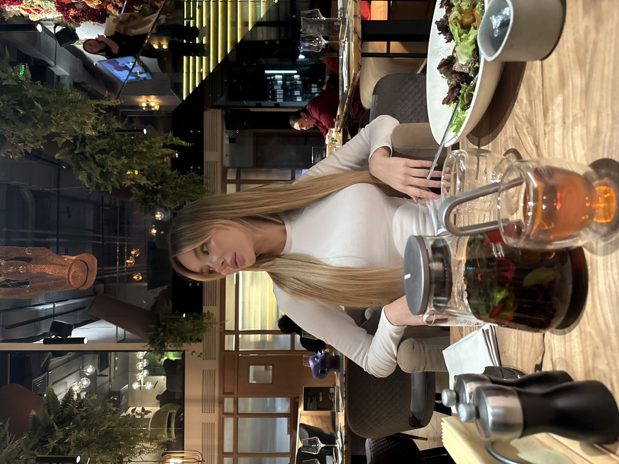 The image shows a young woman with long blonde hair sitting at a table in a restaurant in Moscow, Russia. She is wearing a white long-sleeved shirt. On the table, there is a salad, a teapot with tea, and a salt and pepper shaker. A restaurant worker is visible in the background. The restaurant has an elegant interior with plants and decorative elements. A mirrored ceiling is visible.