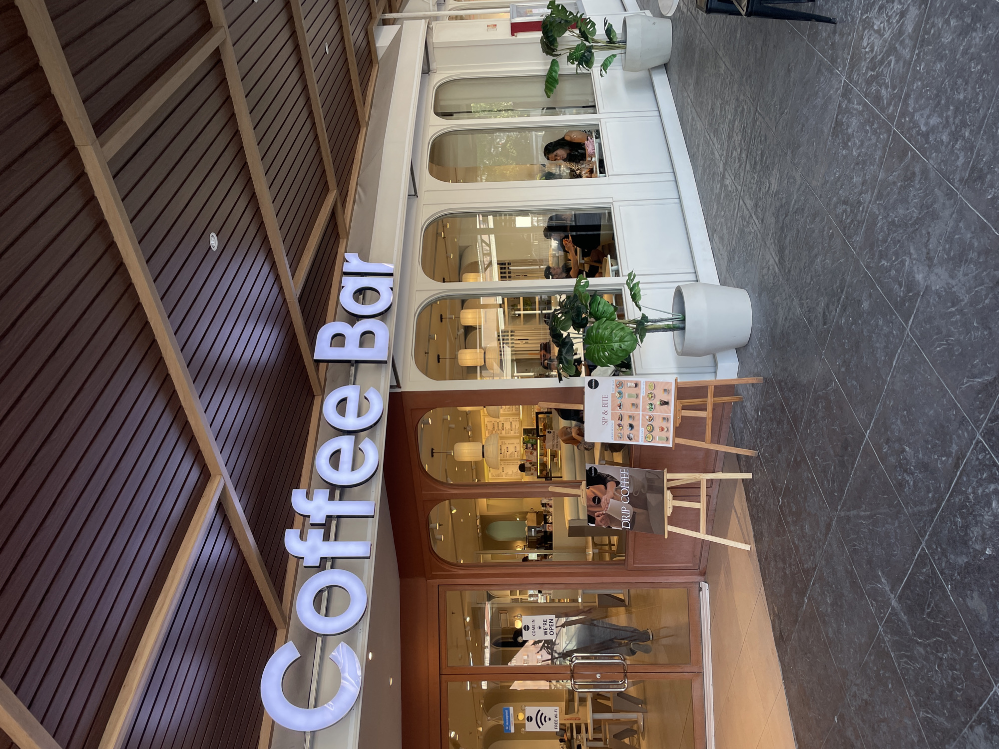 The image shows the exterior of a coffee shop labeled "Coffee Bar". The shop has a modern design with large glass windows and doors, and a wooden slatted ceiling. Inside, patrons can be seen seated at tables, and a barista is working behind the counter. There are also several potted plants outside the coffee shop, adding a touch of greenery to the scene. In front of the entrance, there are two easels displaying menus. The coffee shop is located in Davao City, Philippines, as indicated by the presence of a "Free Wi-Fi" sign with the Globe Telecom logo, a prominent telecommunications company in the Philippines. The overall atmosphere appears to be casual and inviting, suggesting a popular spot for coffee and relaxation.