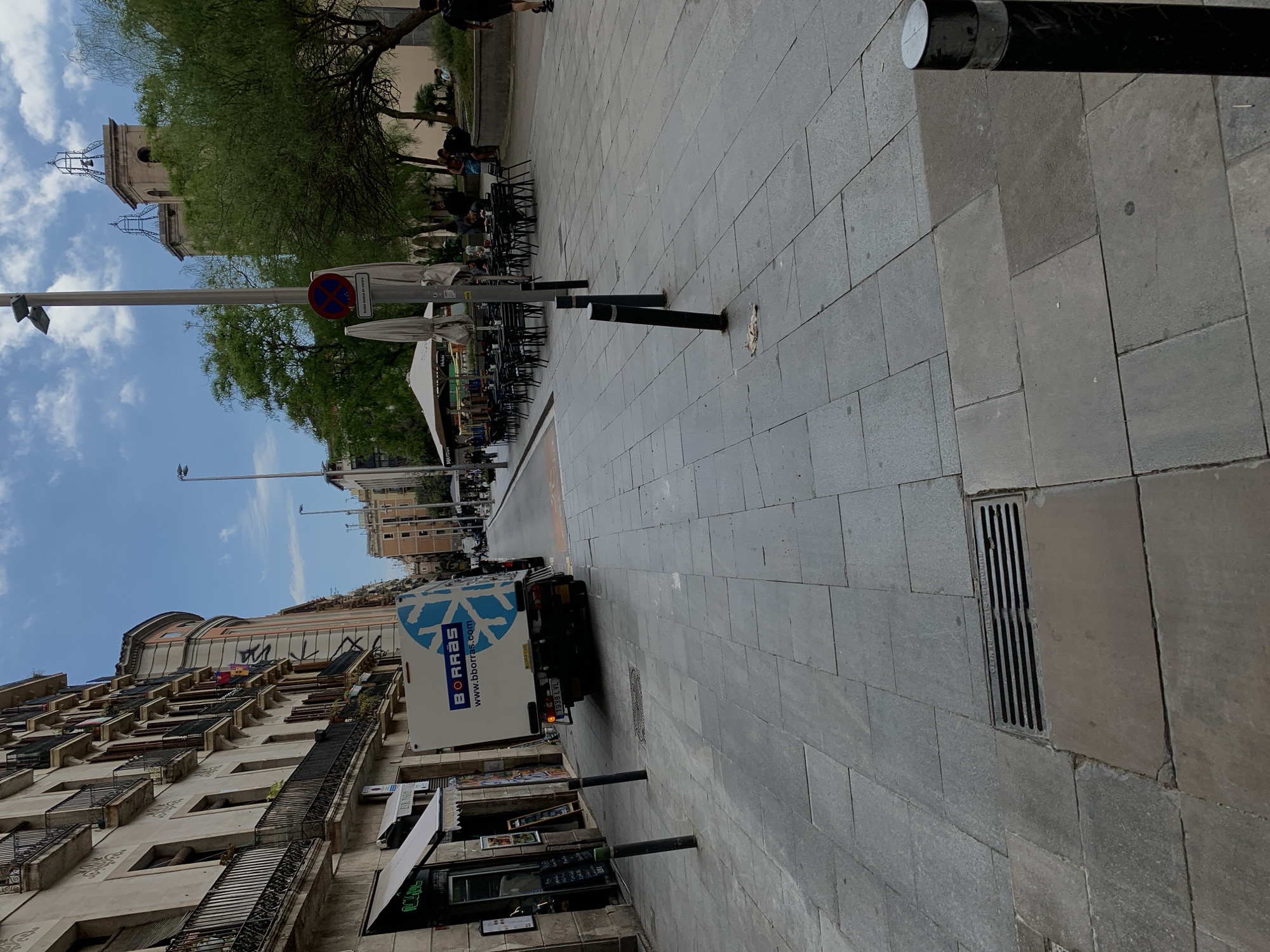 A street in Barcelona, Spain. There are multi-story buildings with balconies on the left side of the street. A white truck with the word "BORRAS" on its side is driving down the street. Tables and chairs are set up outside of a restaurant or cafe on the right side of the street. A tower and church can be seen in the distance. The street is paved with gray stone blocks. A no-entry road sign is also visible. Some trees are along the street.