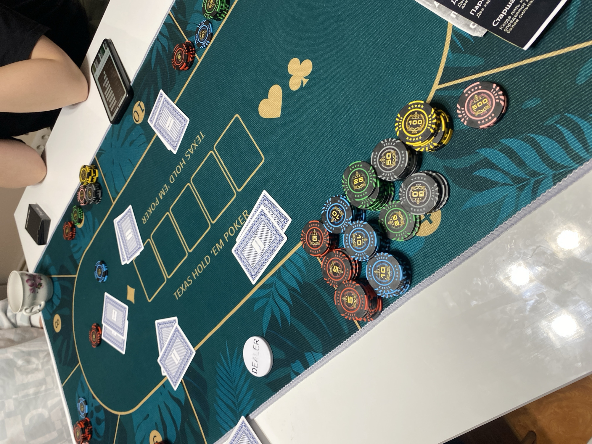 The image shows a poker game in progress, specifically Texas Hold 'em, with the game board and chips arranged on a green felt-covered table decorated with leaf designs and card suit symbols. The table is positioned against a white surface, likely a table or countertop.
Several stacks of poker chips are present, including denominations of 5, 10, 25, 50, 100, and 500. There is also a "DEALER" button. The community cards are laid out, and player cards are visible in front of each betting position.
A woman is partially visible in the upper left, sitting at the table. A phone and a mug are also present on the table.
The text on the table cloth reads "TEXAS HOLD 'EM POKER", which indicates the game being played. There is also a card laying on the table with text in Russian, referencing poker strategies.
Based on the presence of Russian text and information available about the image, it's implied to be in Chaikovsky, Russia.