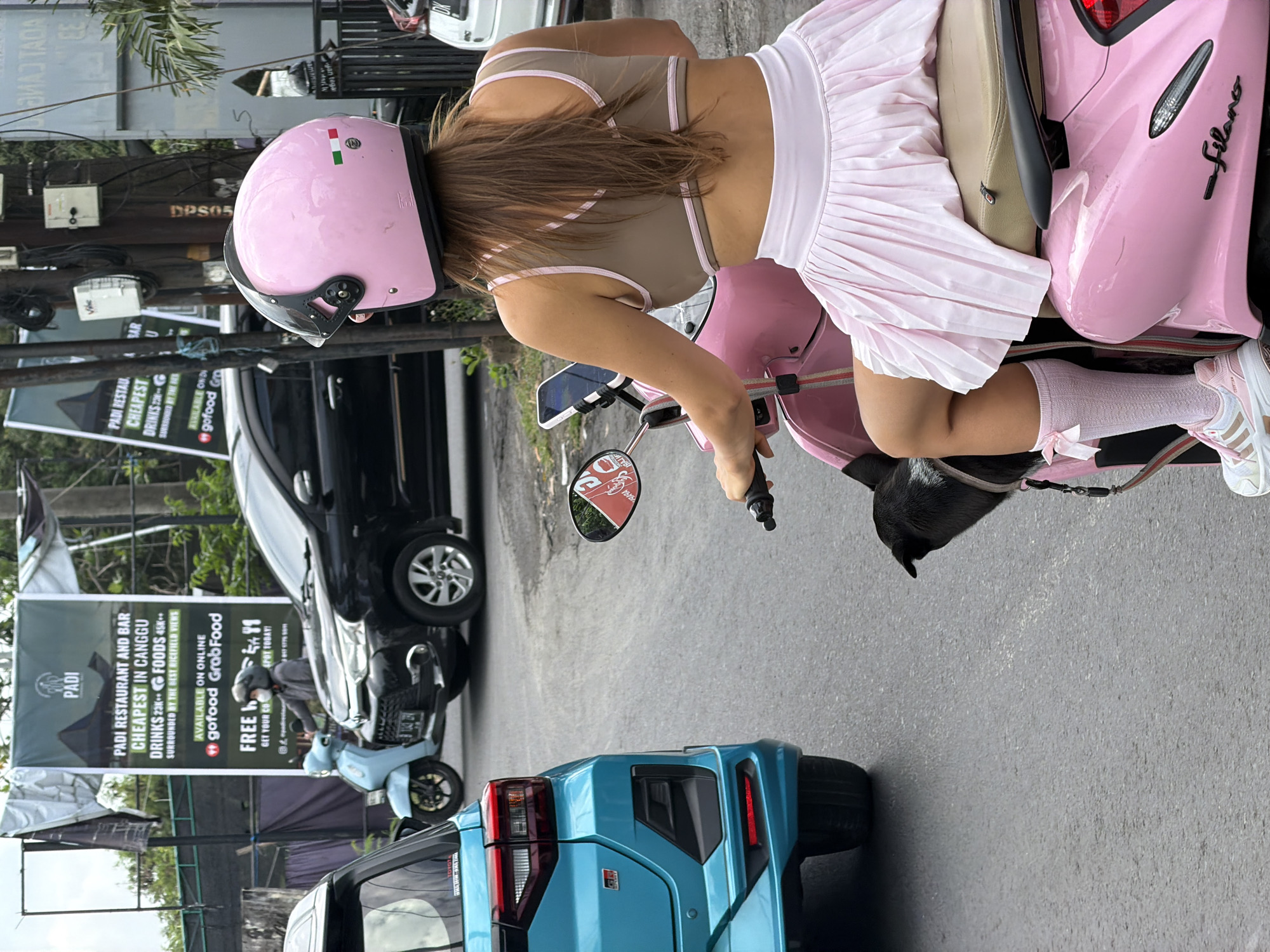 In North Kuta, Indonesia, a woman wearing a pink helmet, a light brown and pink top, and a pink pleated skirt is riding a pink scooter. A small black dog is perched on the back of the scooter. The woman is looking ahead, and a mobile phone is mounted on the scooter's handlebars. In the background, a turquoise car is visible, along with other vehicles and a large banner for "PADI RESTAURANT AND BAR". The banner advertises "CHEAPEST IN CANGGU" for drinks and food, and mentions availability on GoFood and GrabFood. The scene is set on a paved road, likely a street in a populated area.